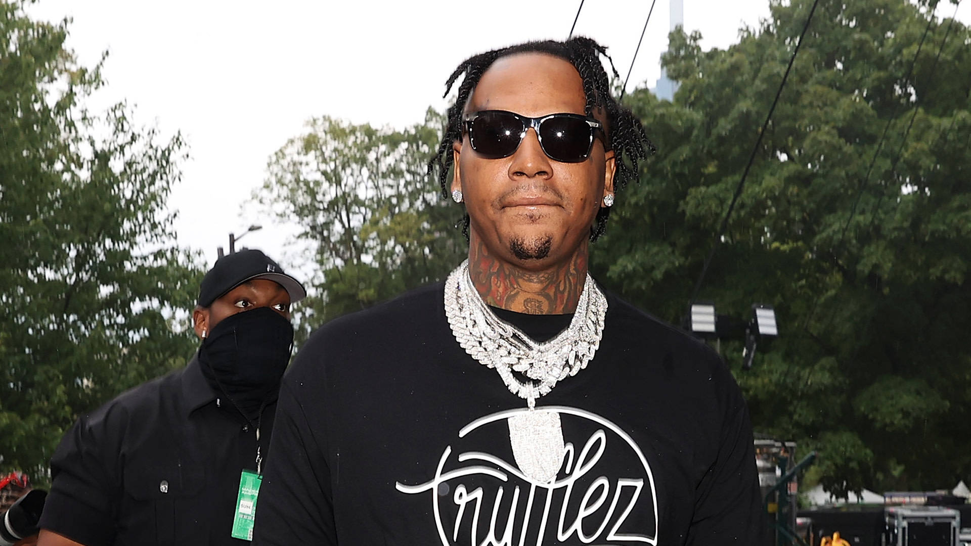 時尚 Moneybagg Yo 饒舌歌手 壁紙 — 一個留著雷鬼辮、戴著太陽眼鏡的男人，穿著黑色T恤和厚重的銀鏈，站在戶外，身後有樹木。