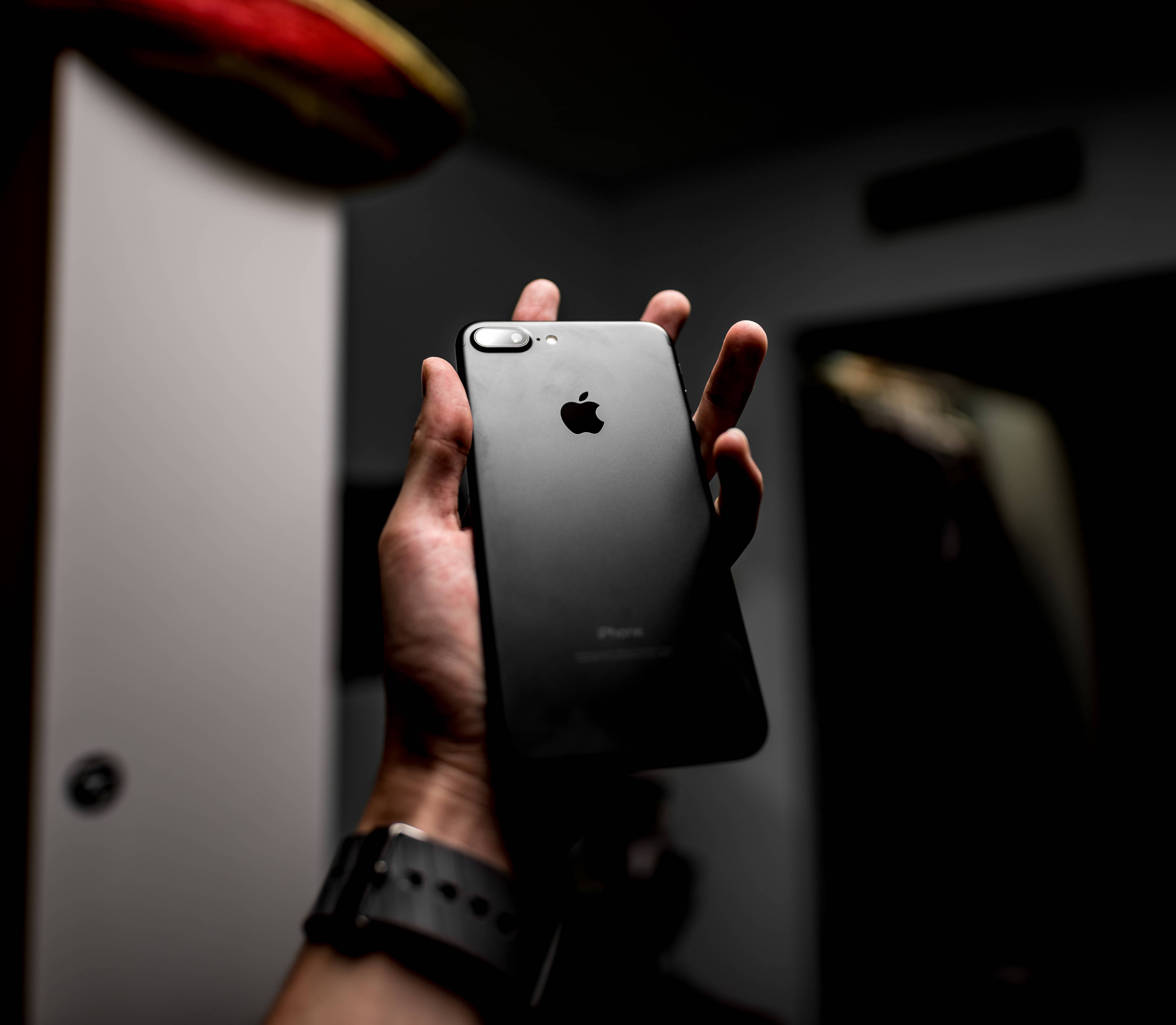 時尚iPhone 8搭配吸睛背景 壁紙 — 一隻手拿著帶有Apple標誌的黑色iPhone，背景是暗色模糊的柔和燈光。