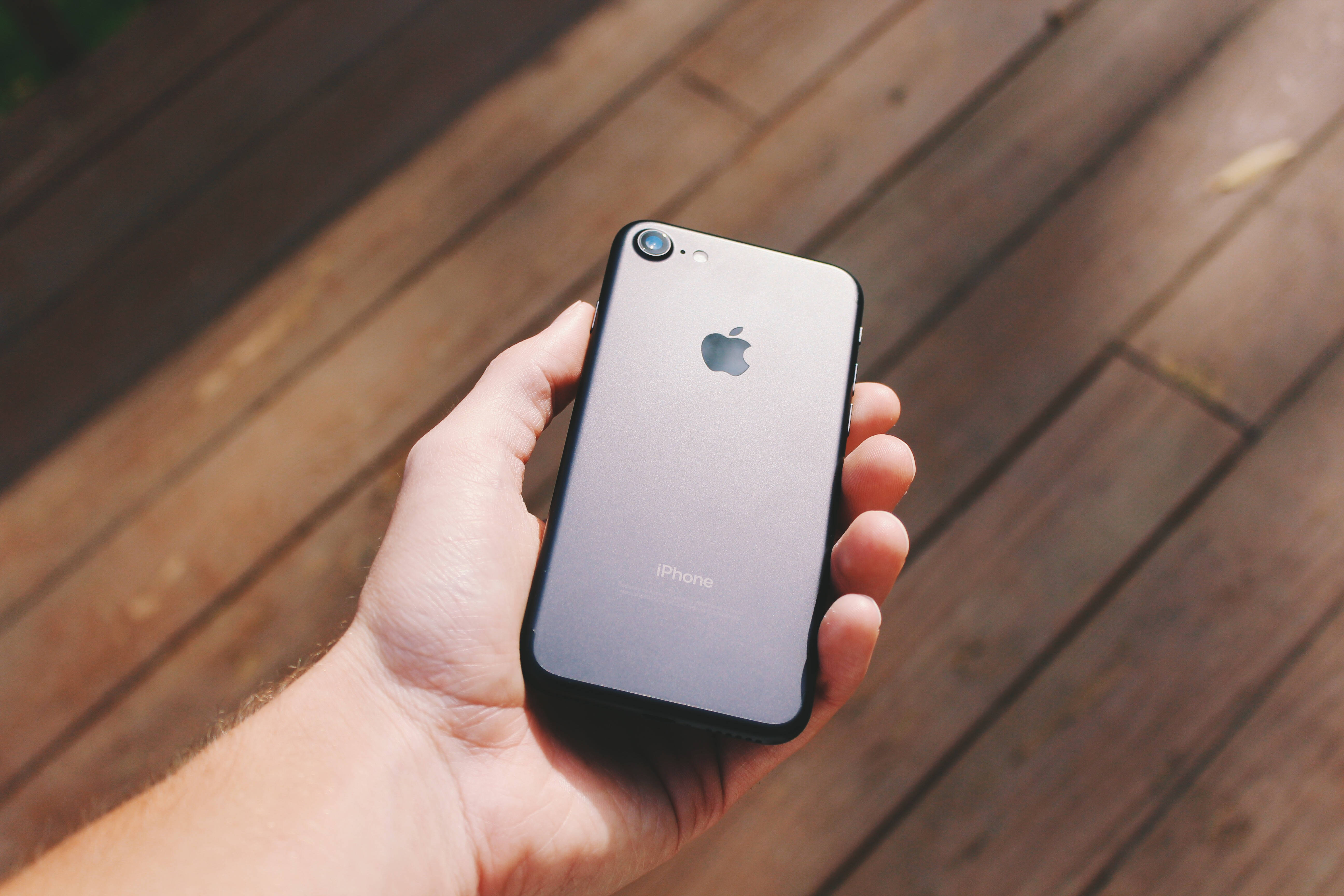 時尚iPhone 8搭配壯麗背景 壁紙 — 一隻手在明亮的陽光下，在溫暖的木質甲板上拿著銀色iPhone 7。