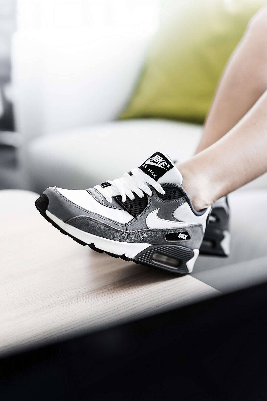 時尚Nike Air Max運動鞋 壁紙 — 一個人腳部嘅特寫，穿著灰色同白色嘅Nike Air Max波鞋，放喺木質表面上，背景係模糊嘅梳化。