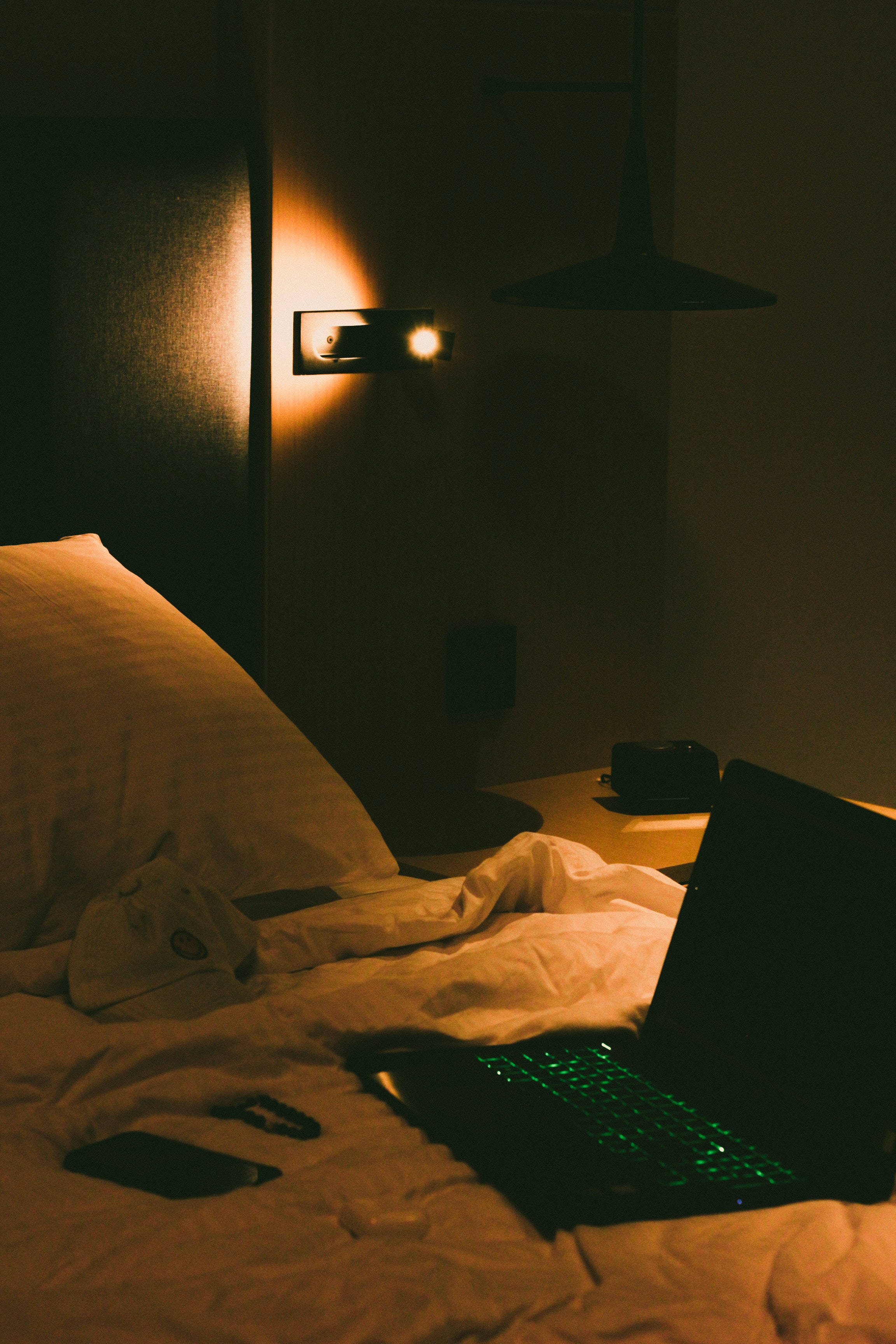 時尚Tumblr筆記本電腦溫馨臥室氛圍 壁紙 — 夜晚昏暗的臥室，發光的筆記型電腦放在皺巴巴的床單上，溫暖的床頭燈投下柔和陰影。