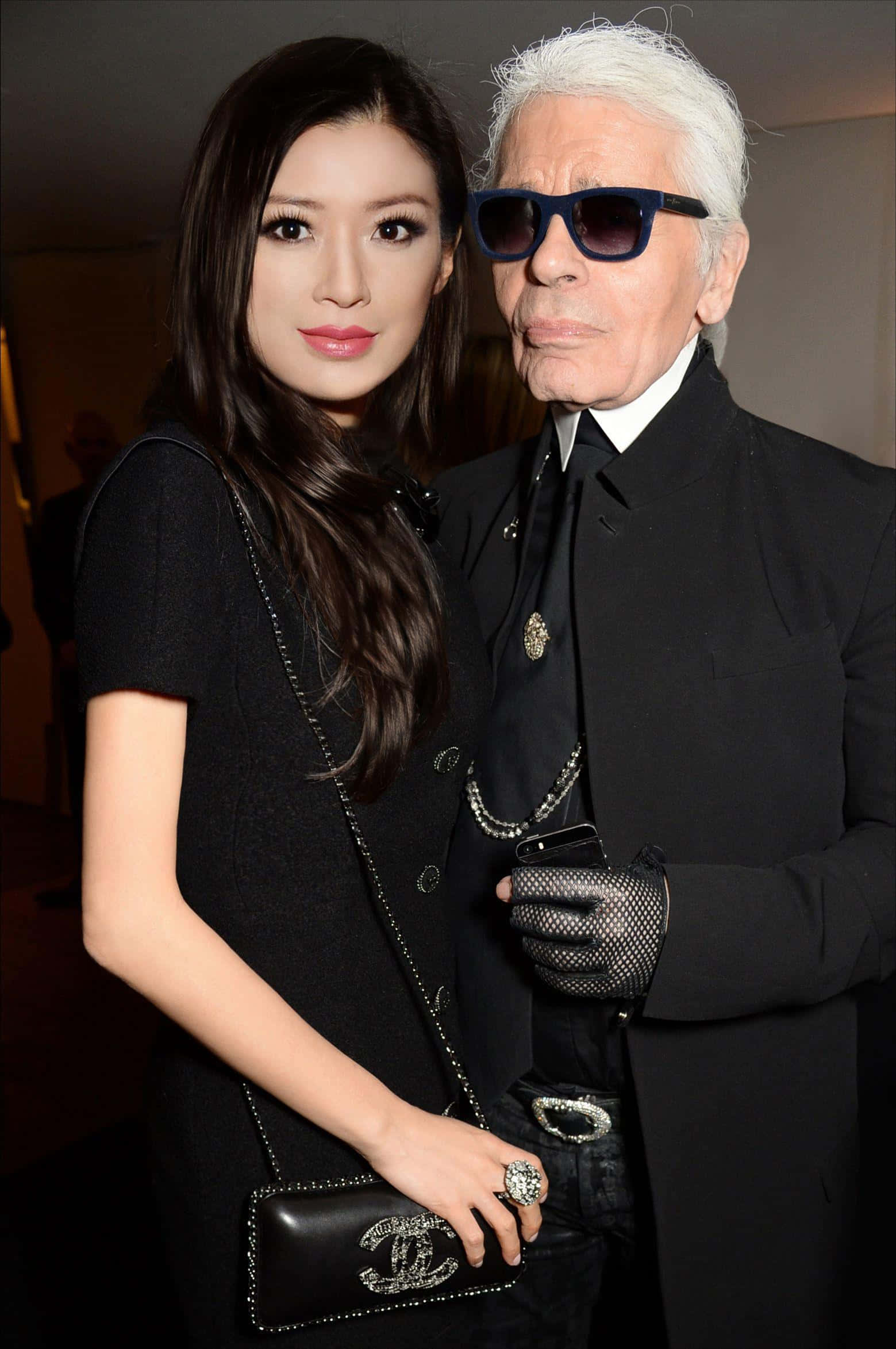 時尚偶像 Karl Lagerfeld 壁紙 — 一位長黑髮的女性和一位白髮戴太陽眼鏡的男性在室內活動中穿著黑色正裝一起擺姿勢。