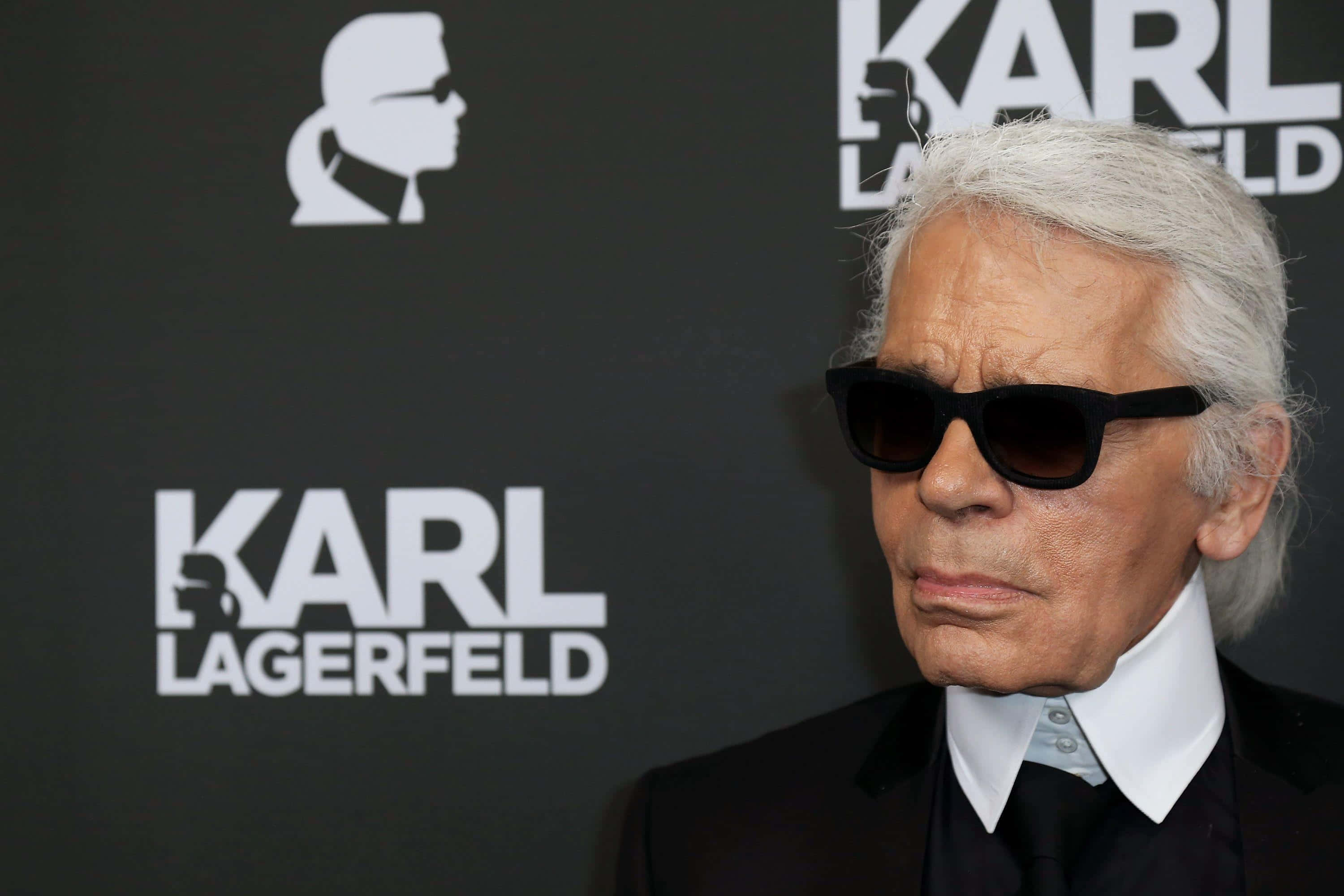 時尚傳奇 Karl Lagerfeld 壁紙 — 卡爾·拉格斐戴著黑色太陽眼鏡和西裝，站在黑色背景前，背景上有他的名字和剪影標誌。