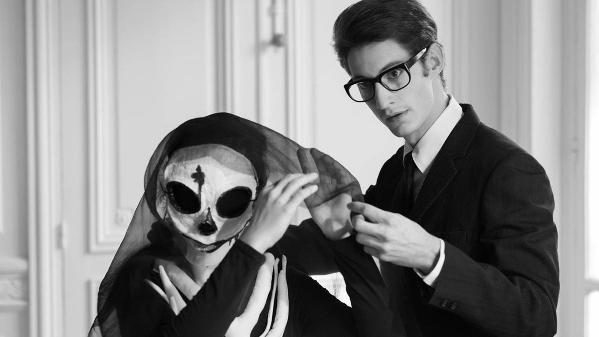 時尚傳奇Yves Saint Laurent 壁紙 — 一名身穿西裝的男子在黑白鮮明的肖像中調整覆蓋在戴著骷髏面具的人身上的面紗。
