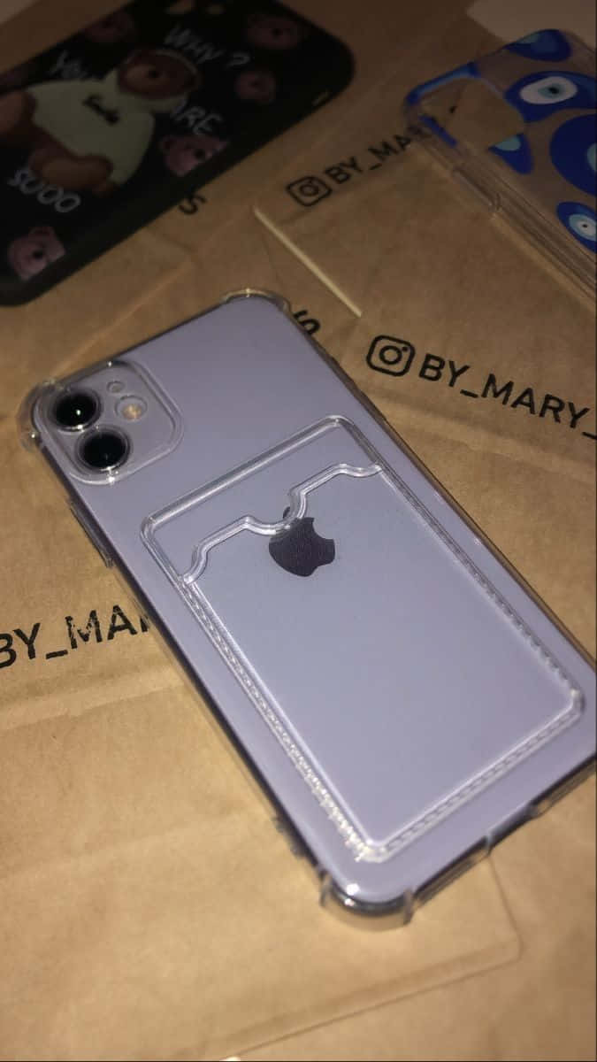 時尚優雅的 iPhone 12，搭配微妙漸層邊框 壁紙 — 淺紫色iPhone放在透明手機殼（帶卡槽）中，擱在印有BY_MARY品牌的棕色紙上。