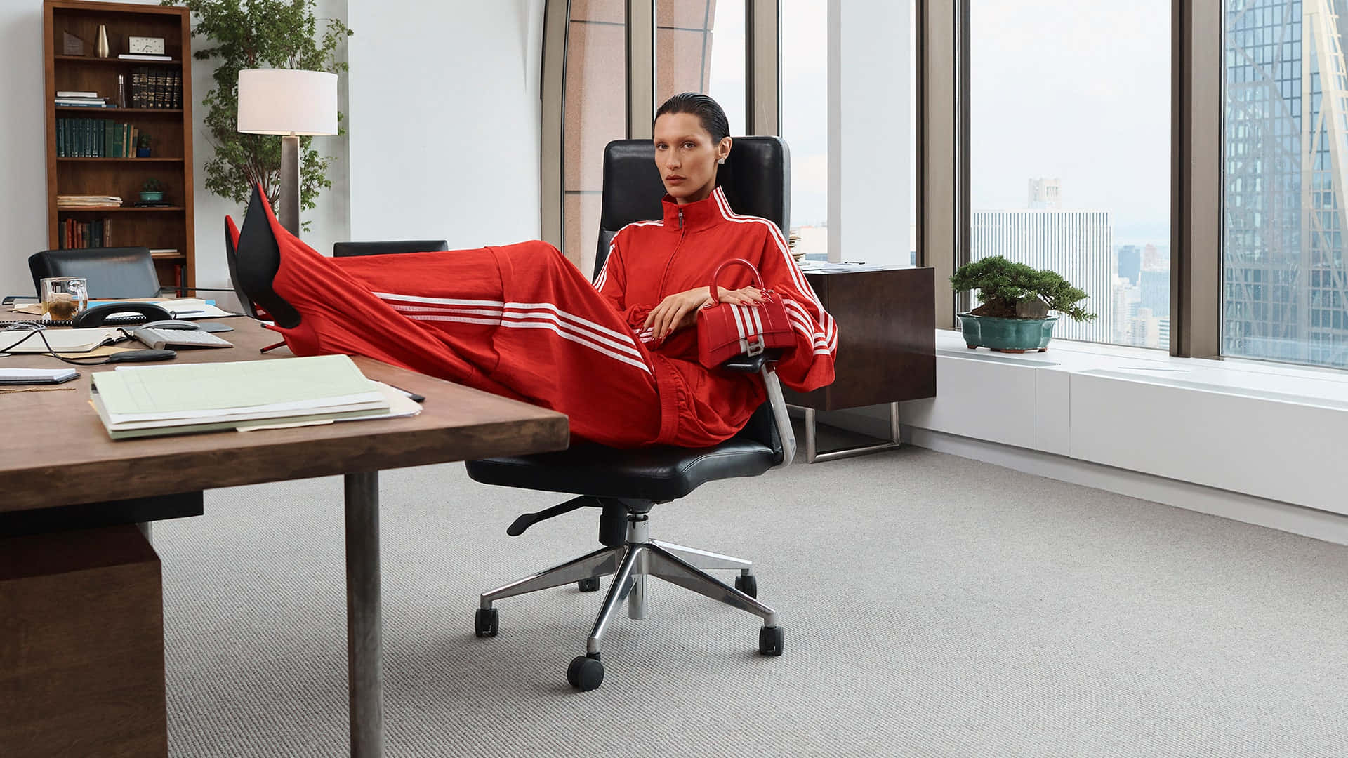 時尚動感 壁紙 — 一位身穿大膽紅色運動服的女性，自信地靠在現代辦公椅上，透過大窗戶欣賞城市景觀。