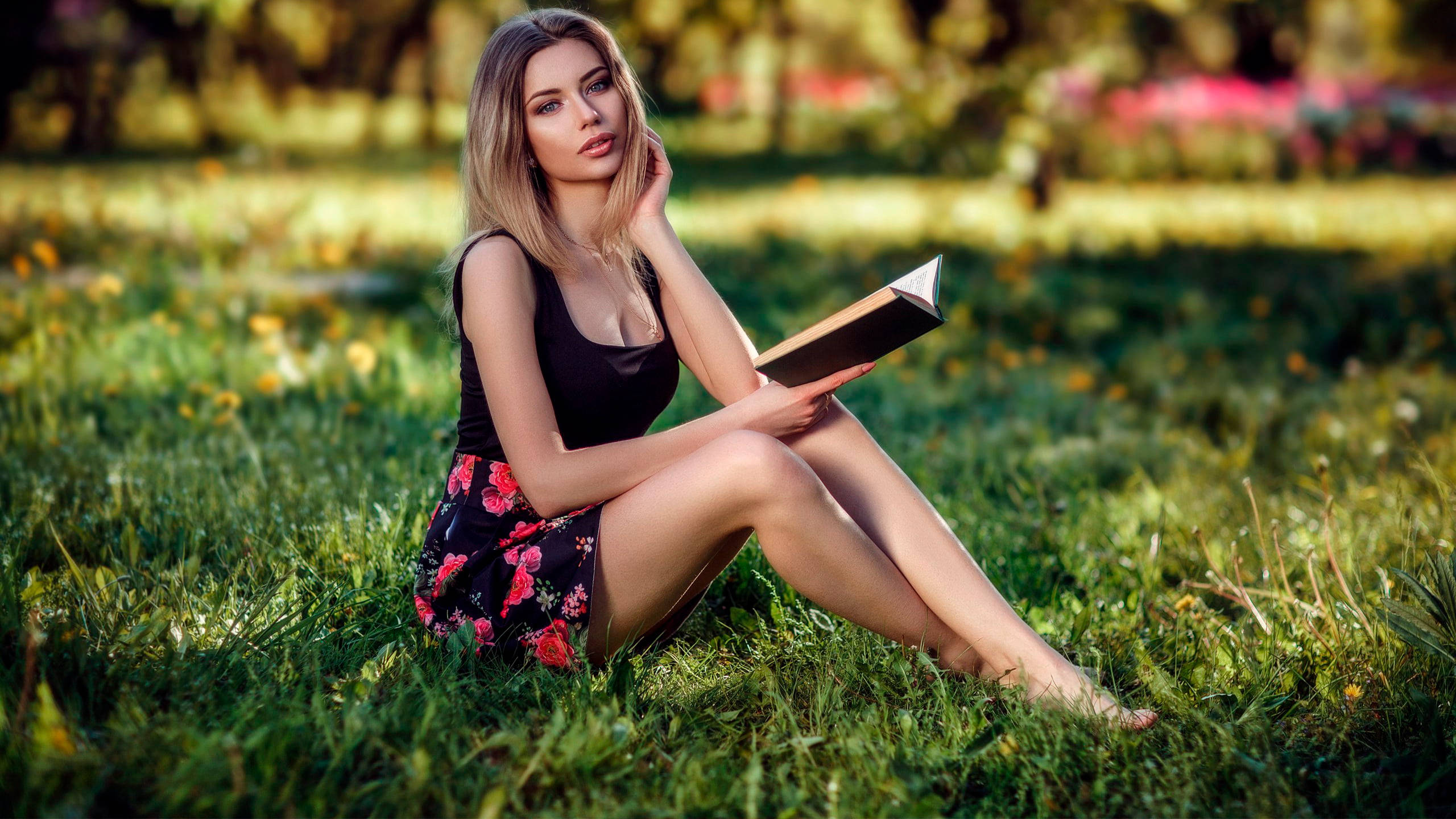 時尚女孩草地讀書 壁紙 — 一位金髮女性赤腳坐在陽光照耀的草地上，手持一本書，身穿黑色上衣和花卉圖案裙子。