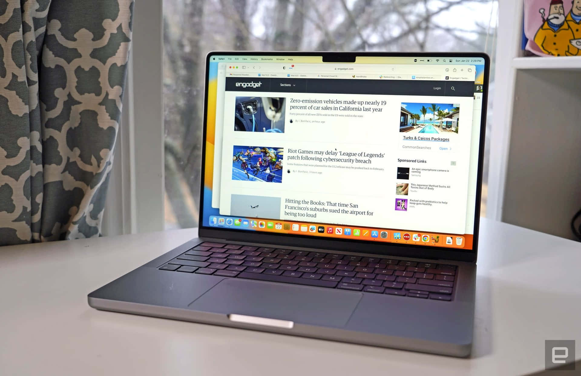 時尚實用，Apple Macbook 提供便利、便攜與強大效能 壁紙 — 一部纖薄的銀色手提電腦打開放在白色桌子上，顯示Engadget新聞頁面，有關於汽車和遊戲的文章，旁邊有圖案窗簾和卡通海報。