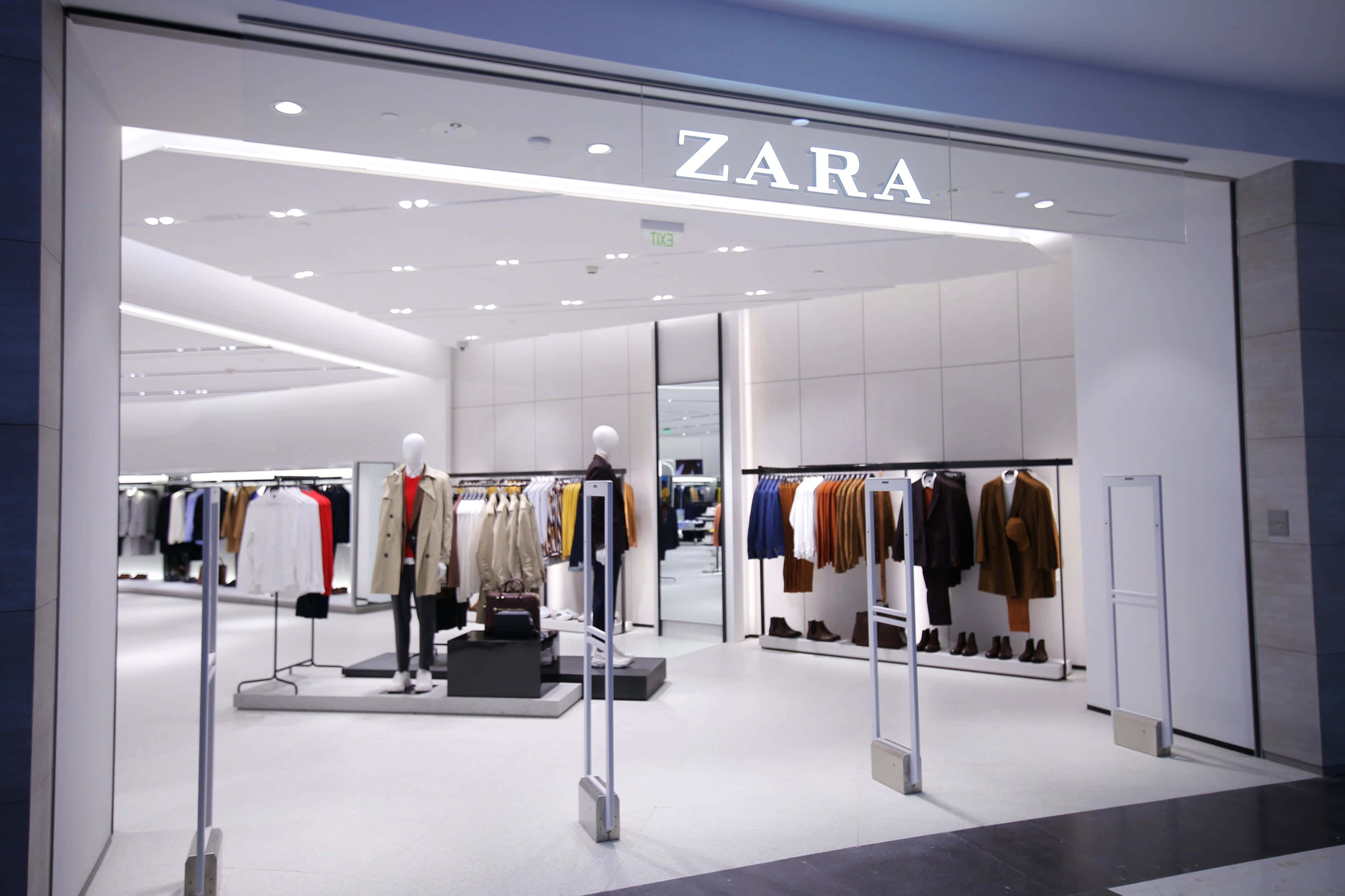 時尚巨擘 Zara 的最新系列 壁紙 — 明亮現代的Zara商店入口，展示整齊排列的衣架和模特兒，在潔淨的白色燈光下。