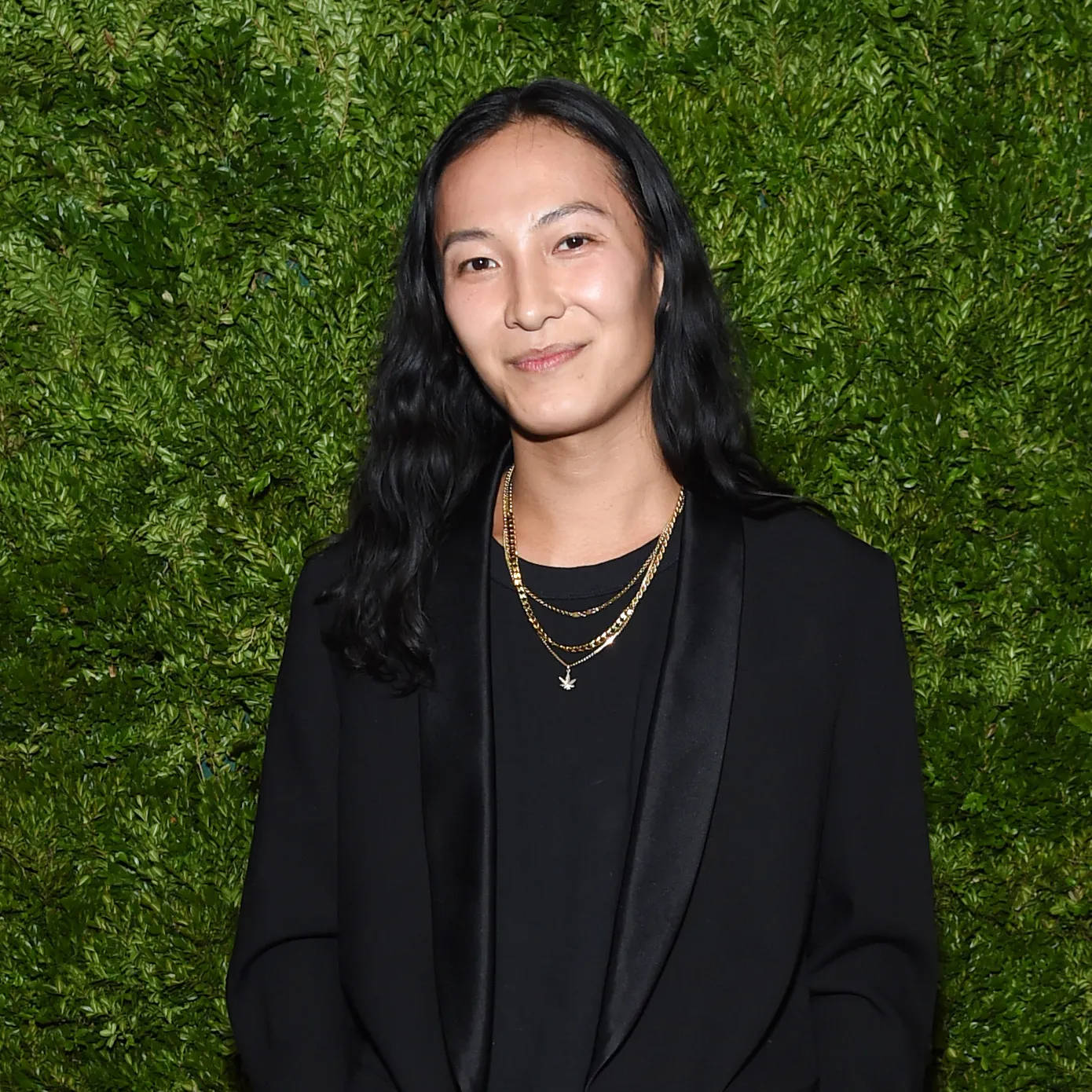身穿黑色西裝外套和金色項鍊的女性對著綠色樹籬牆微笑。