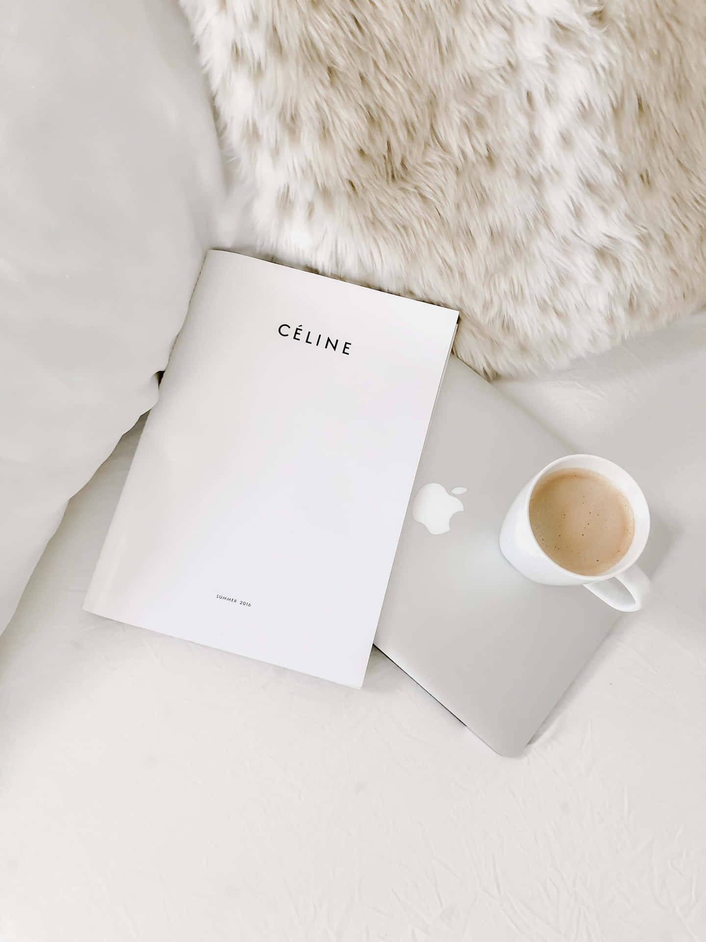 手提電腦、咖啡杯和Celine雜誌放在柔軟的白色紋理床上，配以毛絨絨的米色毯子。