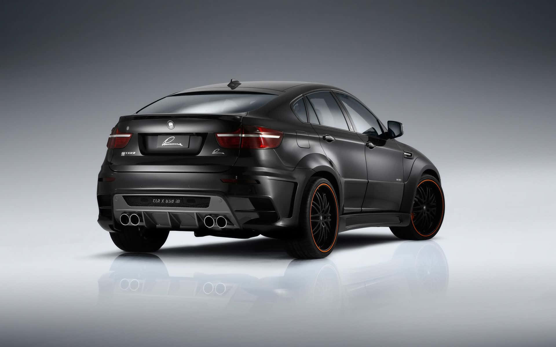 時尚強勁的BMW X6行駛中 壁紙 — 一輛啞光黑色BMW X6M，配備橙色輪圈和四出排氣管，在反射地板的攝影棚中展示。