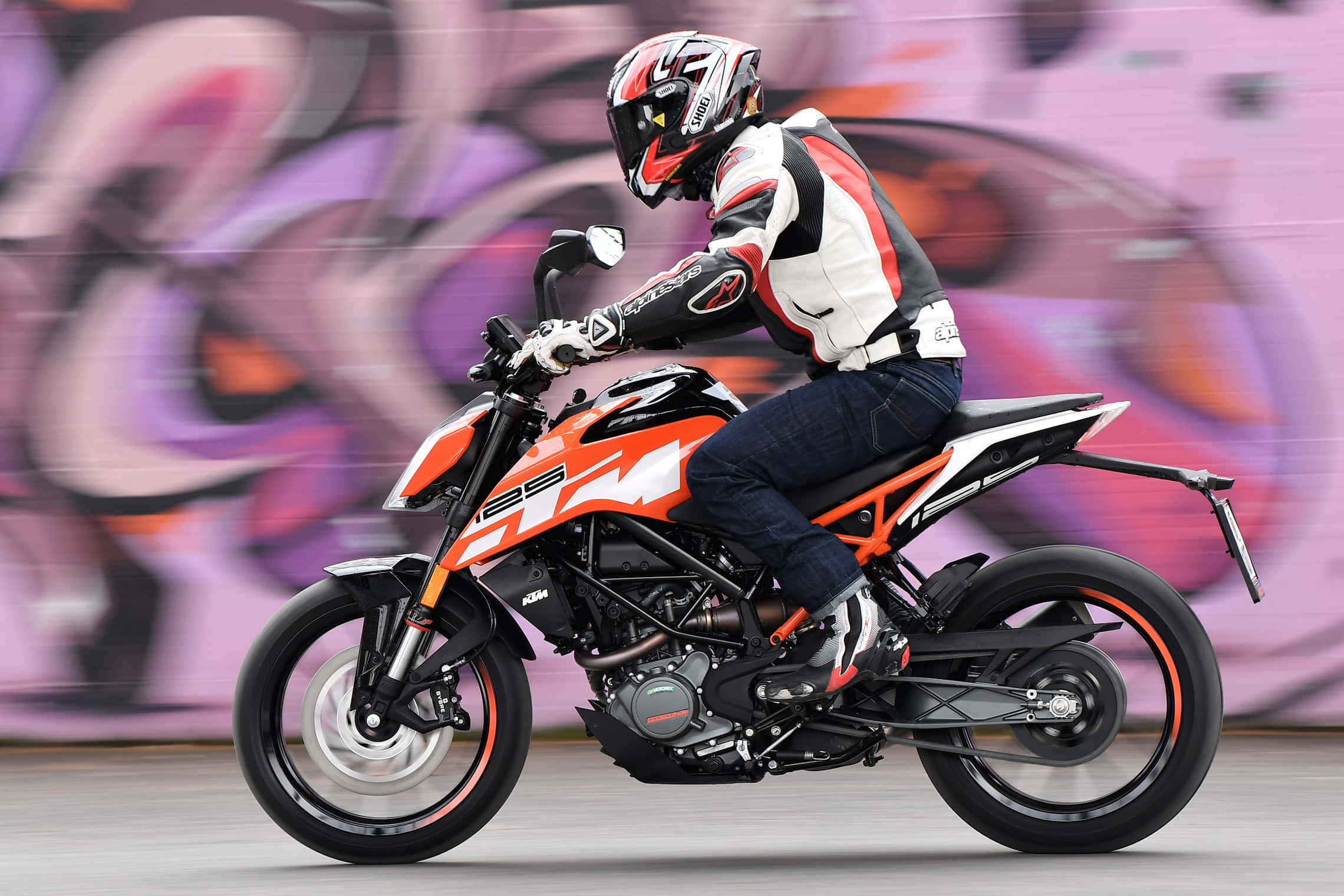 時尚強悍的KTM Duke 壁紙 — 一名全副裝備的電單車手，在充滿活力的塗鴉牆前，騎著橙色KTM電單車，帶有動態模糊效果。
