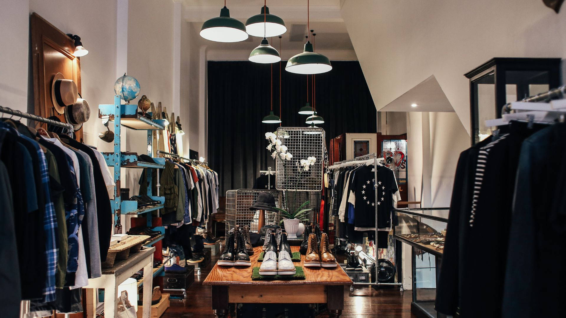 時尚服裝店精品店 壁紙 — 時尚精品店內部，配有吊燈、木製展示桌和衣架，在溫暖的環境照明下。
