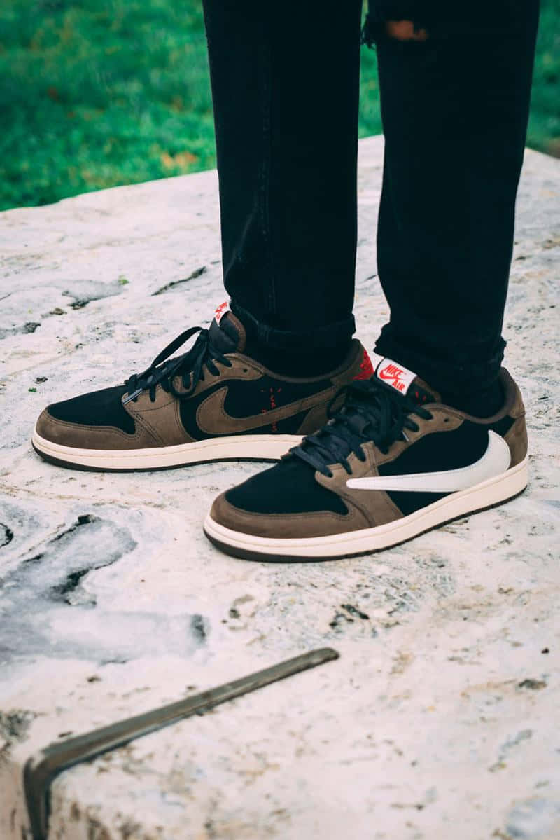 Travis Scott Jordan 1 桌布 壁紙 — 時尚棕色和黑色Nike運動鞋的特寫，放置在風化的石頭表面上，背景有綠色草地。