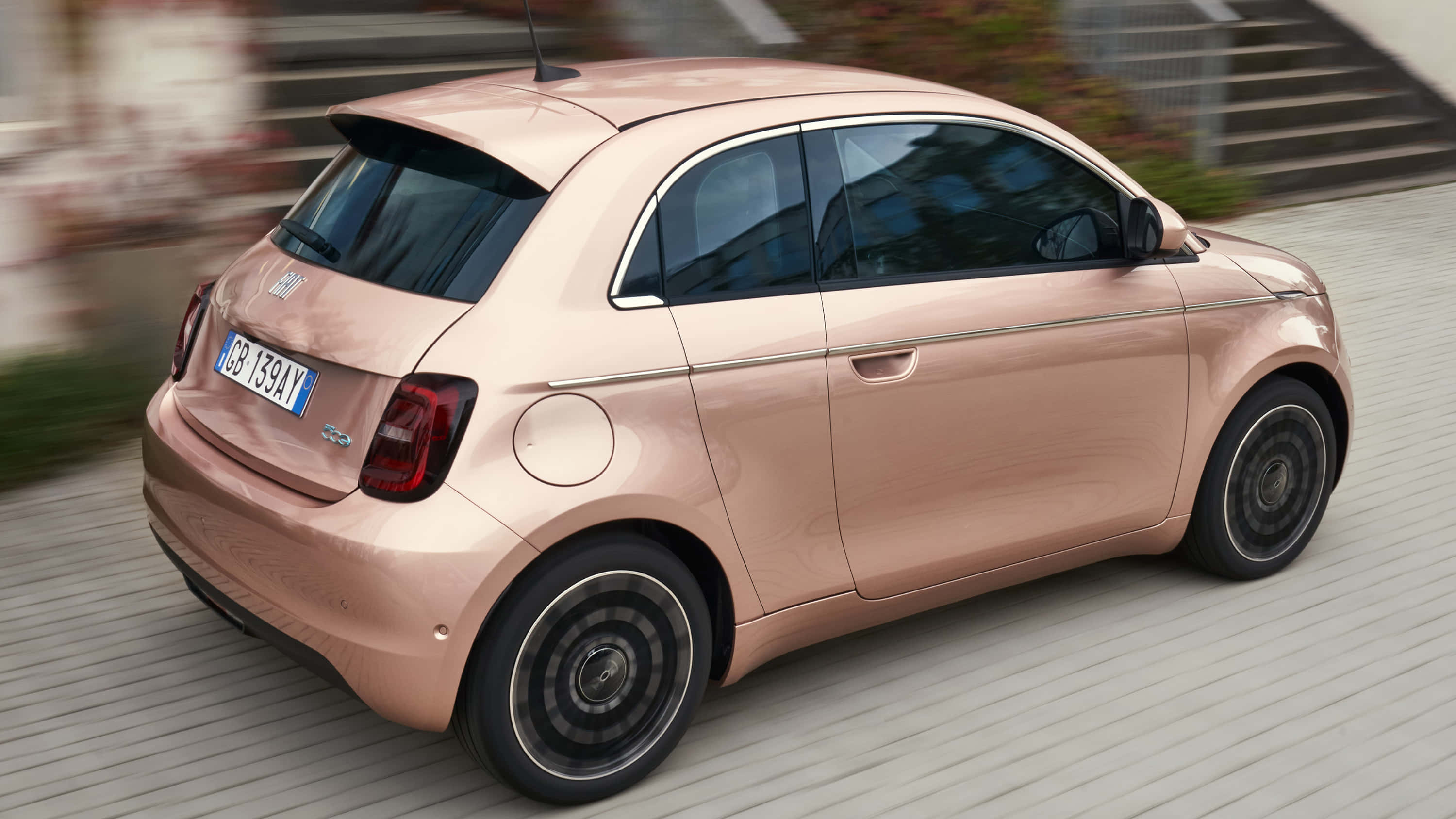 時尚流線型菲亞特500 壁紙 — 一輛玫瑰金色的Fiat 500e電動車在模糊的城市環境中平穩地行駛在木質步道上。