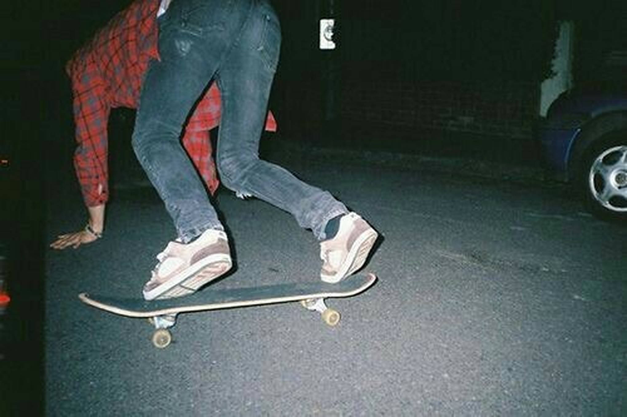時尚滑板男孩夜景 壁紙 — 一個人穿著牛仔褲和格仔襯衫，夜晚在黑暗的街道上蹲低在滑板上，附近有一輛車。