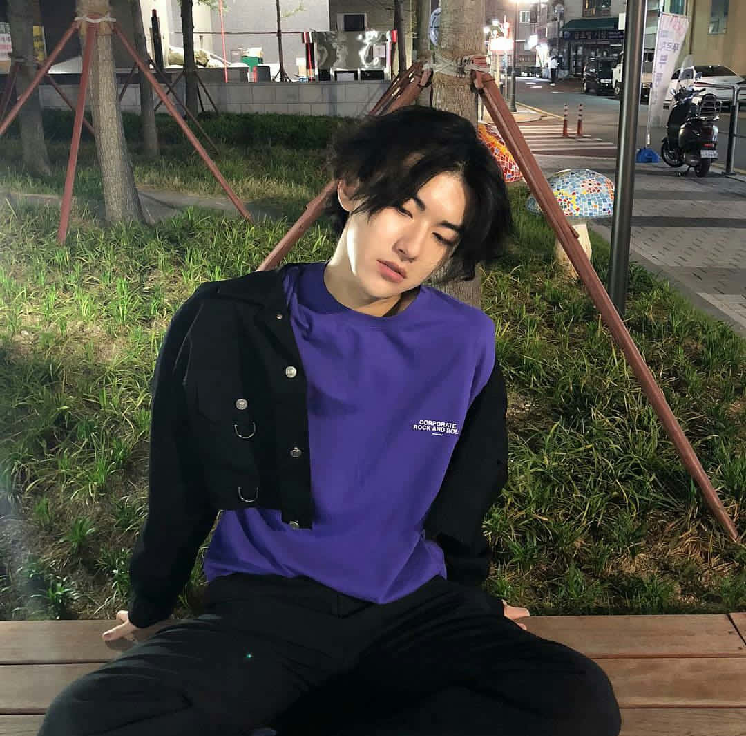 時尚男孩享受美好一天 壁紙 — 一個深色頭髮的年輕男子晚上坐在木長椅上，穿著紫色襯衫和黑色外套，被草地和城市街燈包圍。