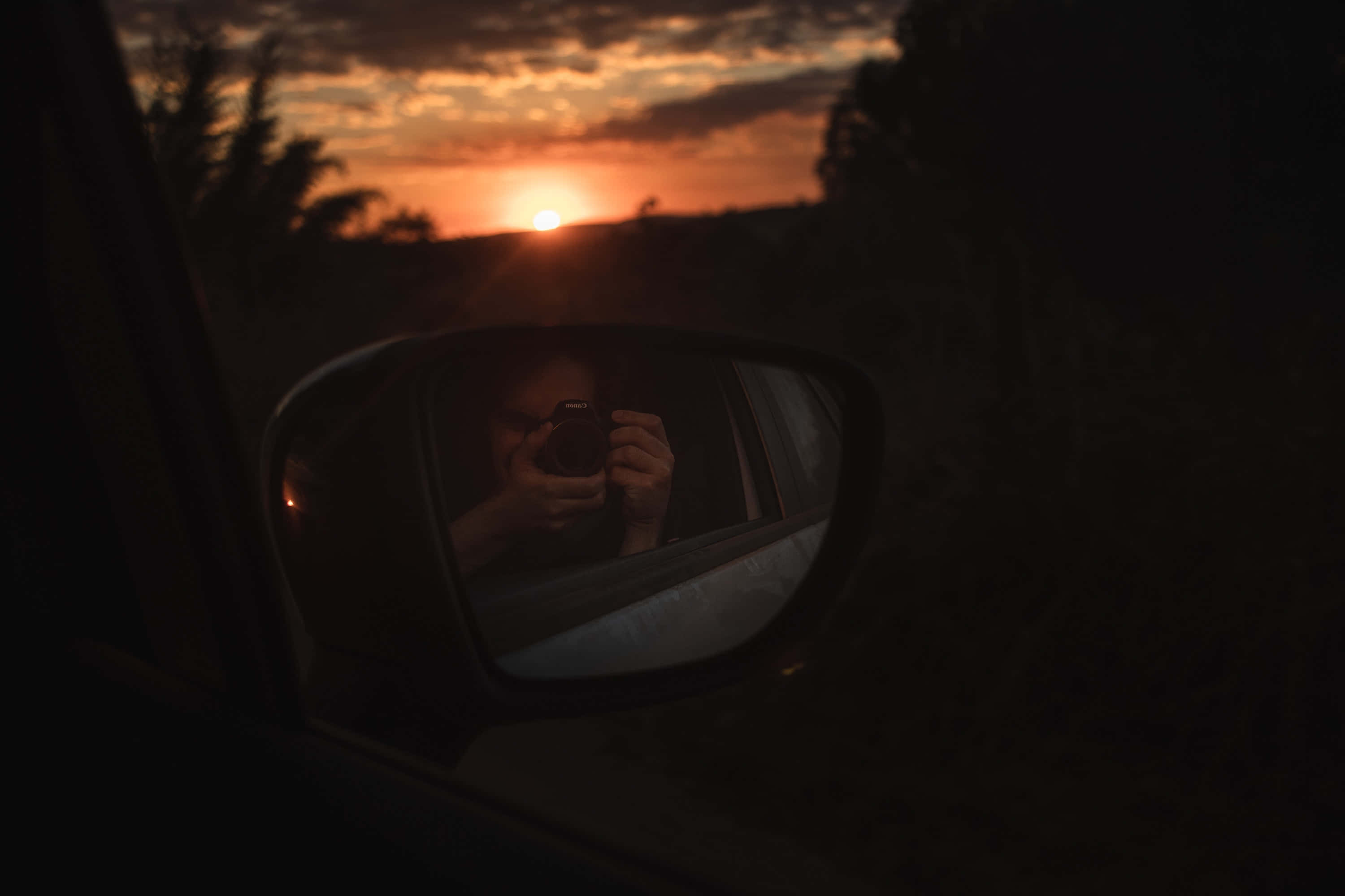 時尚穿搭下的迷人鏡前自拍 壁紙 — 一個人透過汽車側鏡捕捉夕陽，鏡中映出他們的剪影，背景是火紅的橙色雲彩。
