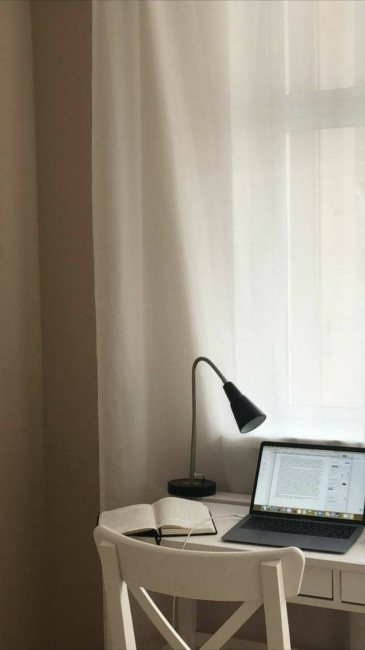白色桌子上有一部打開的筆記型電腦，旁邊有一本書和一個黑色檯燈，柔和窗光照射。