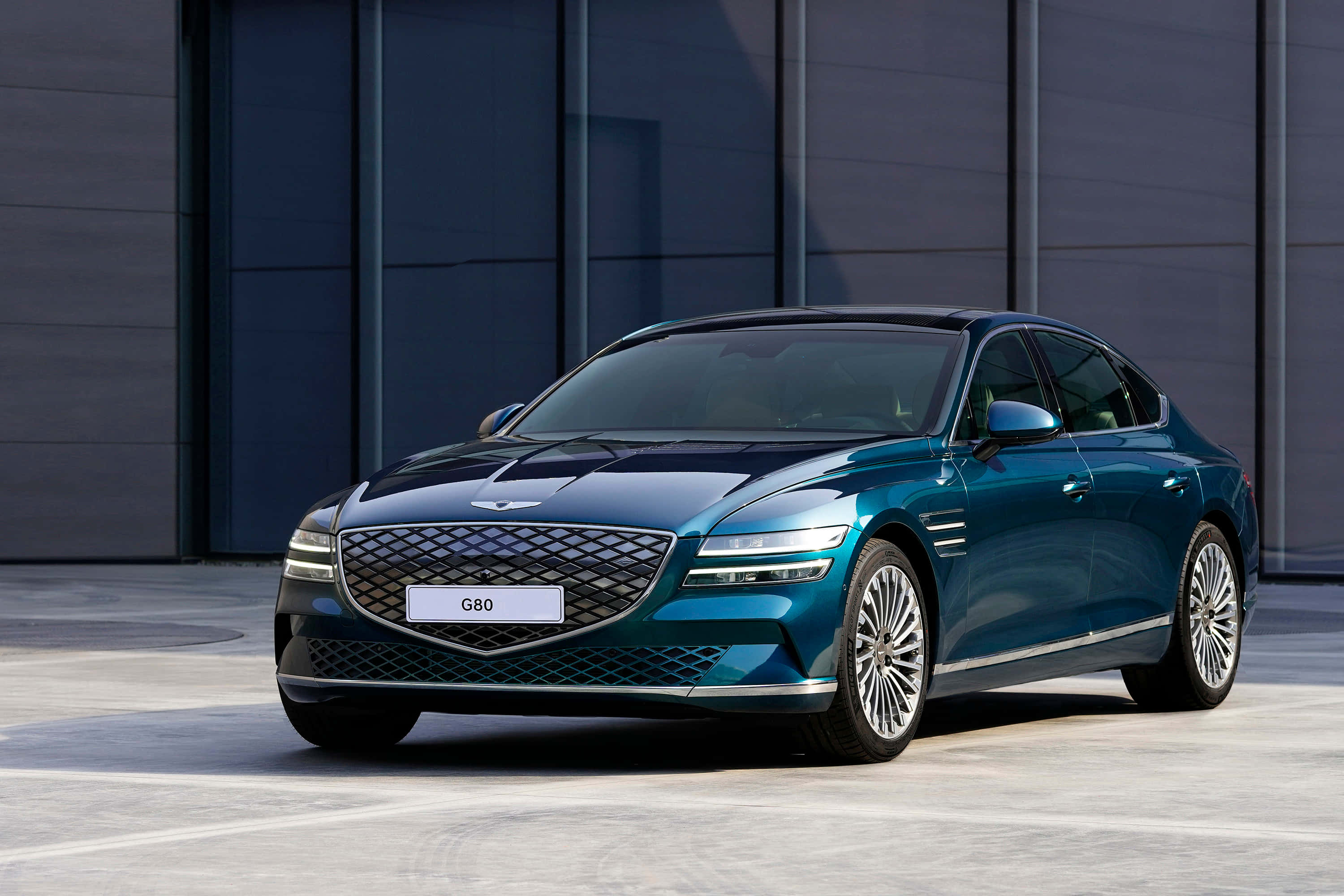 時尚精緻的Genesis G80 壁紙 — 一輛時尚的青色Genesis G80轎車在明亮的日光下，在現代灰色建築外牆前閃閃發光。