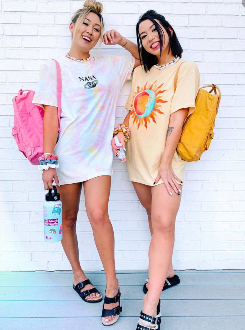兩位穿著休閒夏裝的年輕女性背著背包在白色磚牆前擺姿勢。