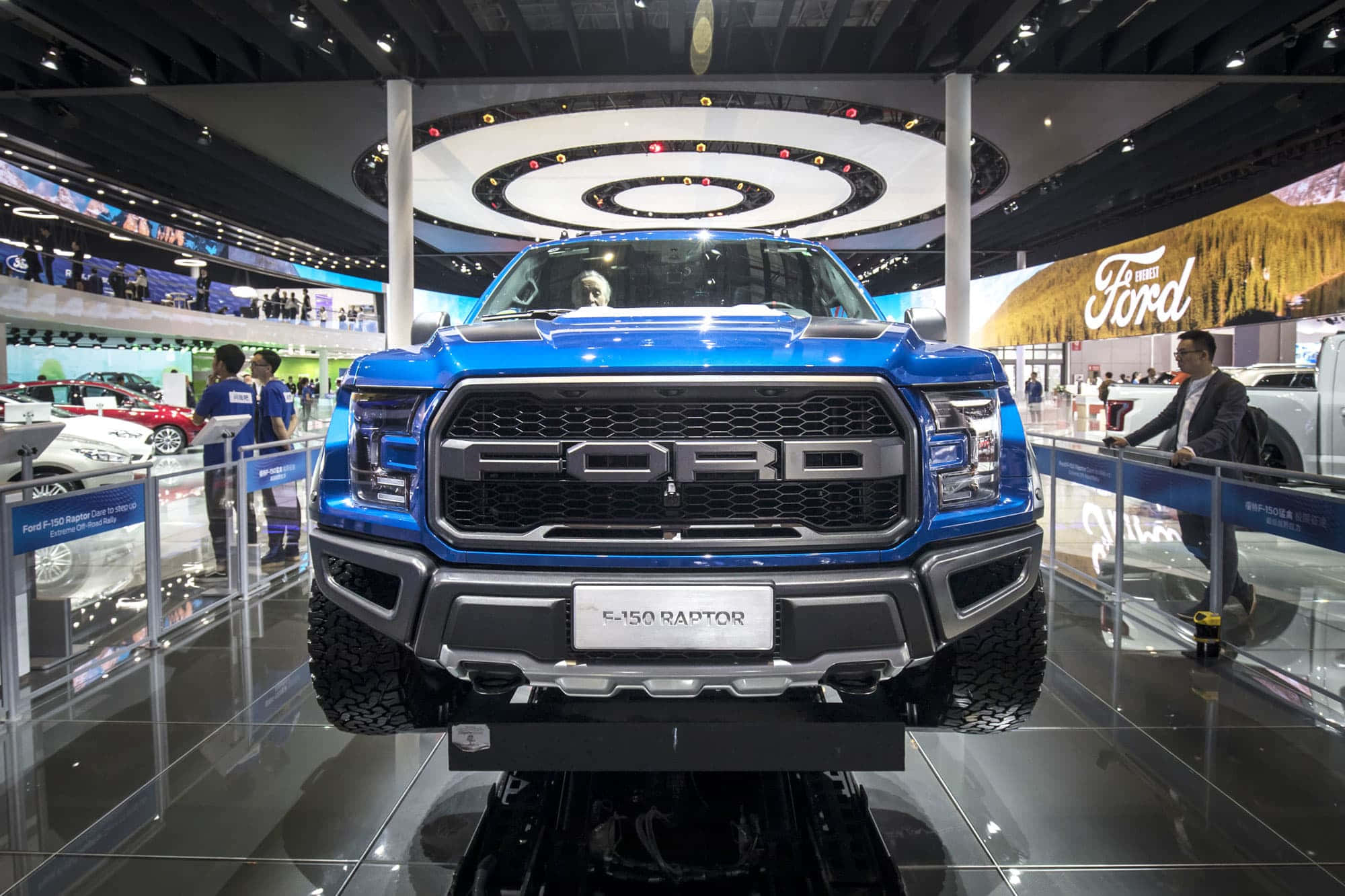 時尚而強勁的福特卡車 壁紙 — 一輛充滿活力的藍色福特F-150 Raptor卡車在繁忙的汽車展覽會上，在明亮的頂燈下展示在光澤地板上。