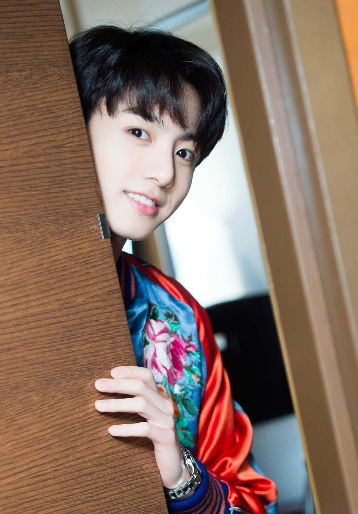 時尚自信的Jungkook 壁紙 — 一個年輕男子穿著鮮豔的花卉緞面夾克，俏皮地從木門後面探出頭來。