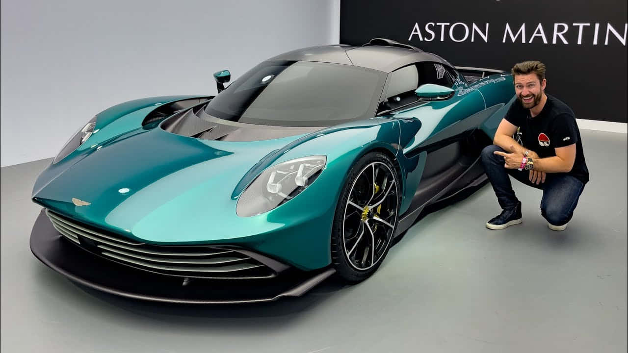 時尚與力量兼具的 Aston Martin 壁紙 — 一名男子蹲在簡約工作室中，旁邊是一輛時尚的青色 Aston Martin 超跑，帶有品牌標誌。