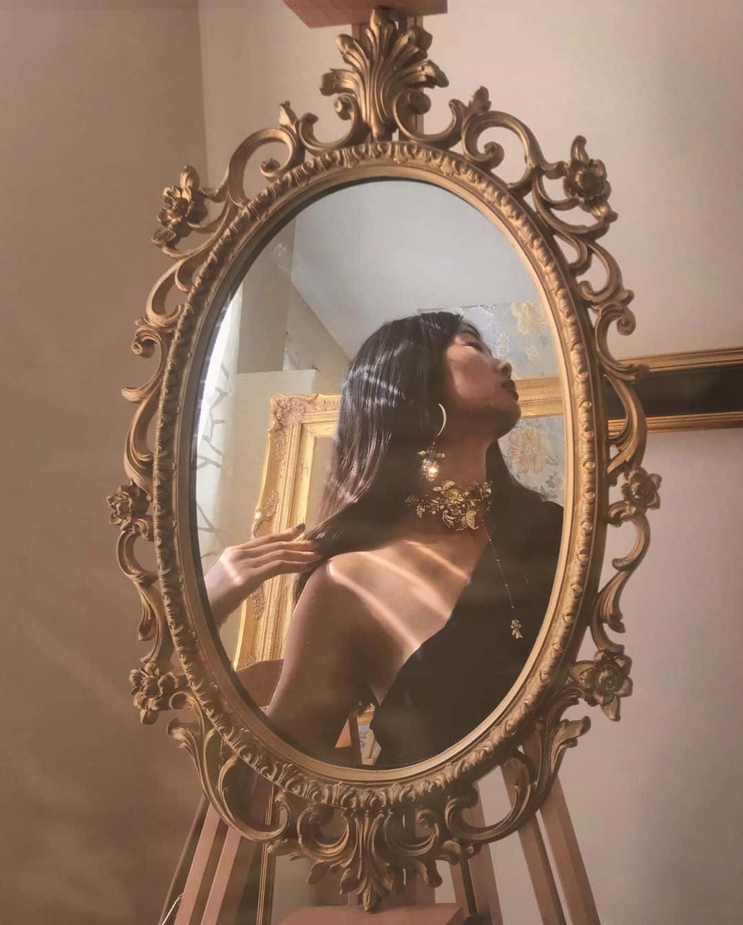 時尚與復古風情 壁紙 — 一位長黑髮的女性抬頭望向一個華麗的金框鏡子，鏡子反射出柔和陽光灑在她的肩膀上。
