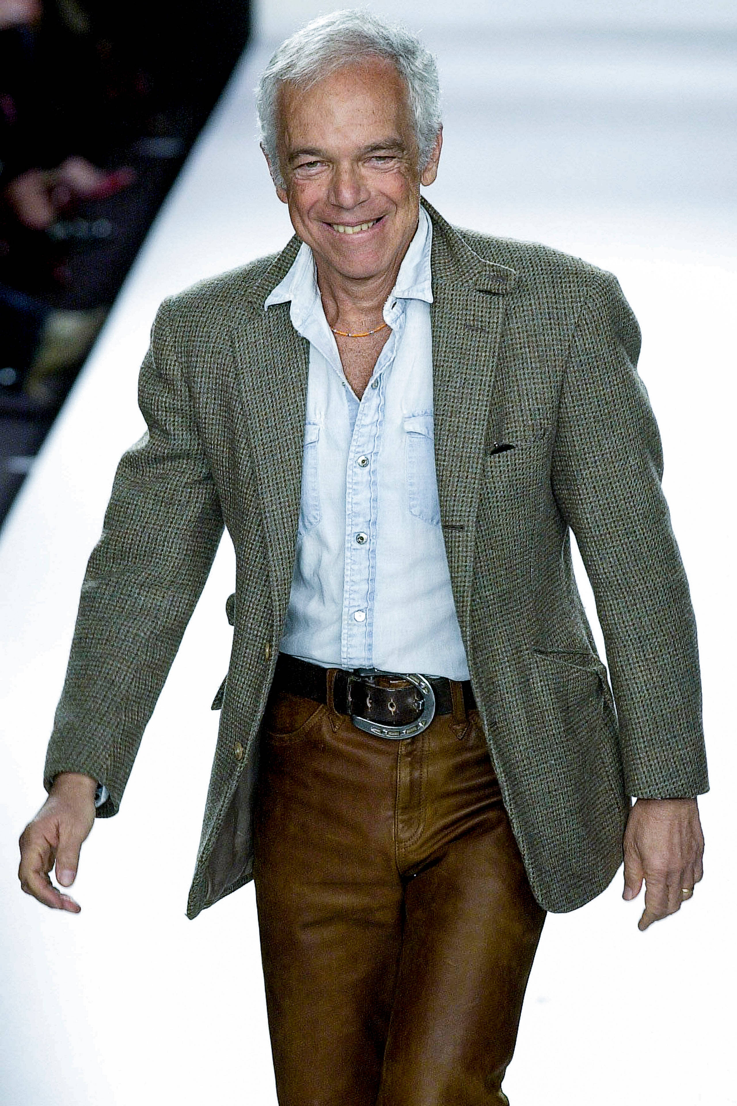 時尚設計師 Ralph Lauren 微笑 壁紙 — 一位微笑的年長男士自信地走在明亮的白色天橋上，身穿綠色格紋西裝外套和棕色皮褲。