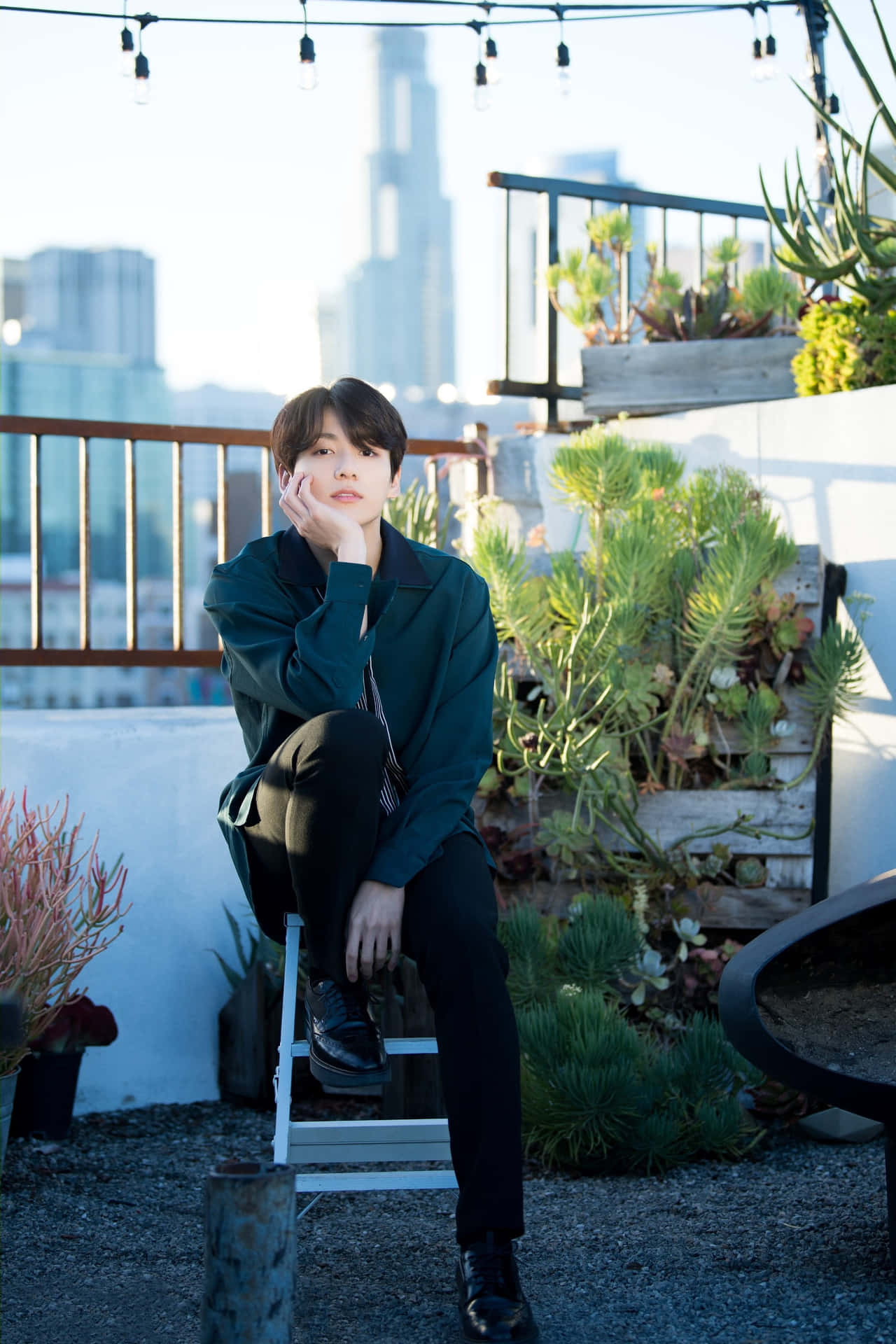 時尚造型的才華洋溢Jungkook 壁紙 — 一名身穿深綠色外套的年輕男子，在陽光普照的屋頂花園中，坐在白色凳子上沉思，背景是城市天際線。