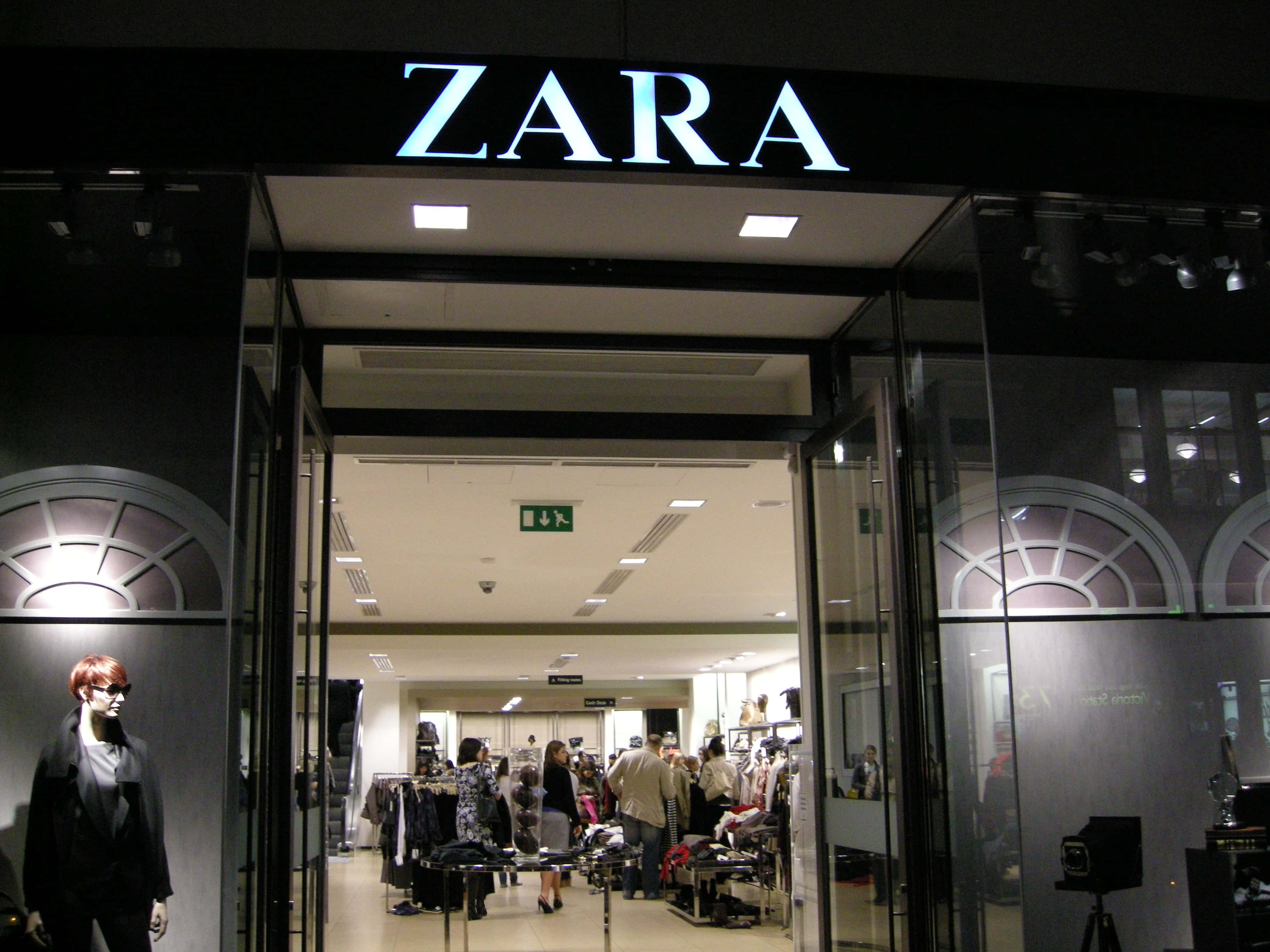 時尚達人展現Zara本季最新造型 壁紙 — 夜晚明亮照明的Zara店舖入口，玻璃門敞開，顧客在店內瀏覽貨架。