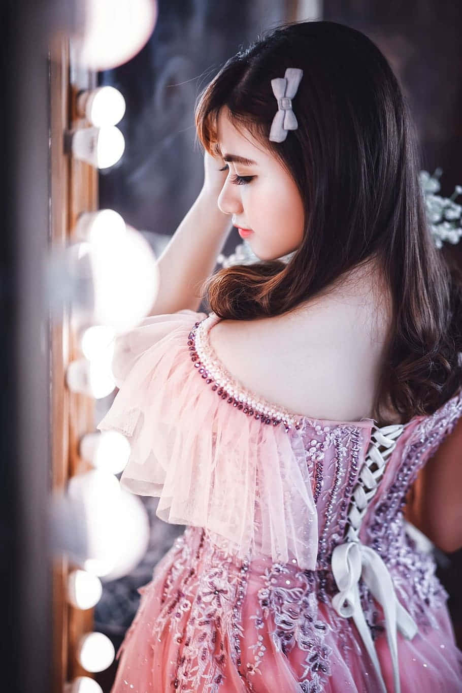 時尚達人穿著優雅晚禮服 壁紙 — 一位身穿粉紅色珠飾長裙的年輕女士，若有所思地凝視著帶有發光燈泡的梳妝鏡。