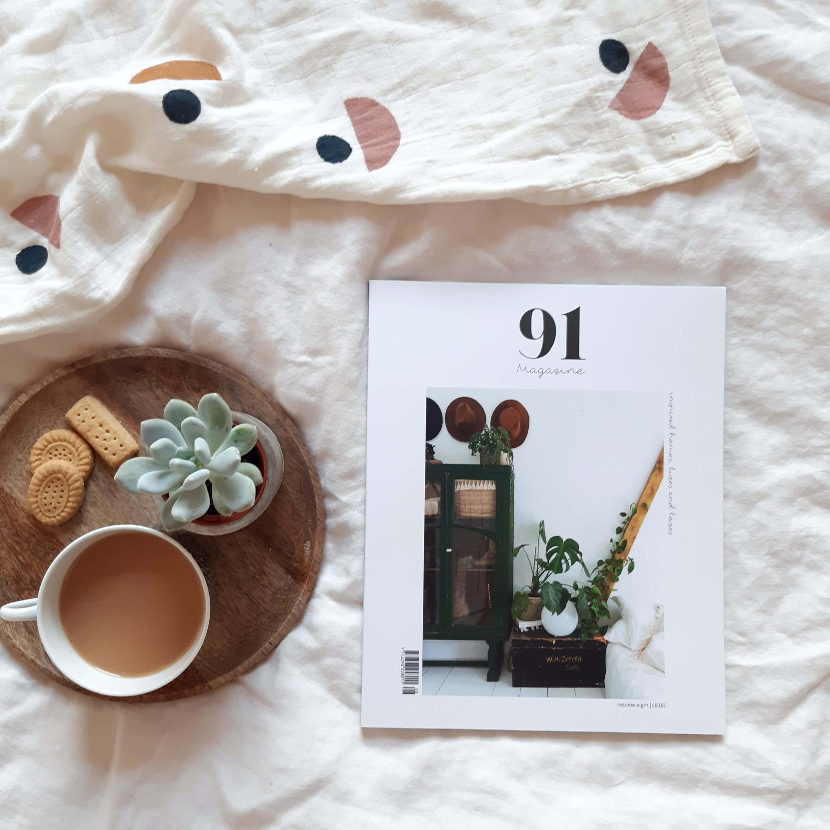 時尚雜誌風向標：音樂、文化、觀點與熱門故事 壁紙 — 一個舒適的平鋪擺設，木托盤上放著茶餅和多肉植物，旁邊有一本雜誌，置於柔軟的白色幾何圖案床單上。
