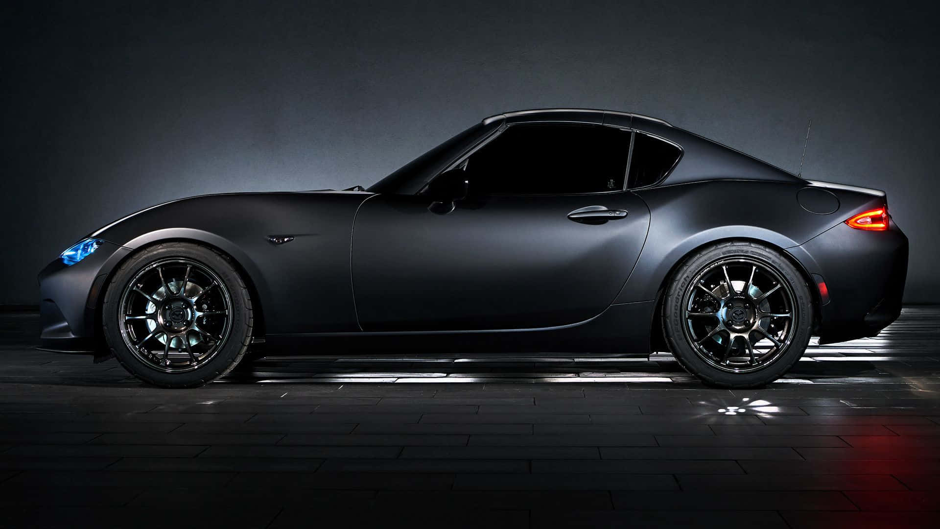 時尚黑色馬自達MX-5 Miata跑車 壁紙 — 一架時尚的啞光黑色跑車，帶有發光的藍色頭燈和紅色尾燈，站在一個帶有戲劇性照明的黑暗工作室中。