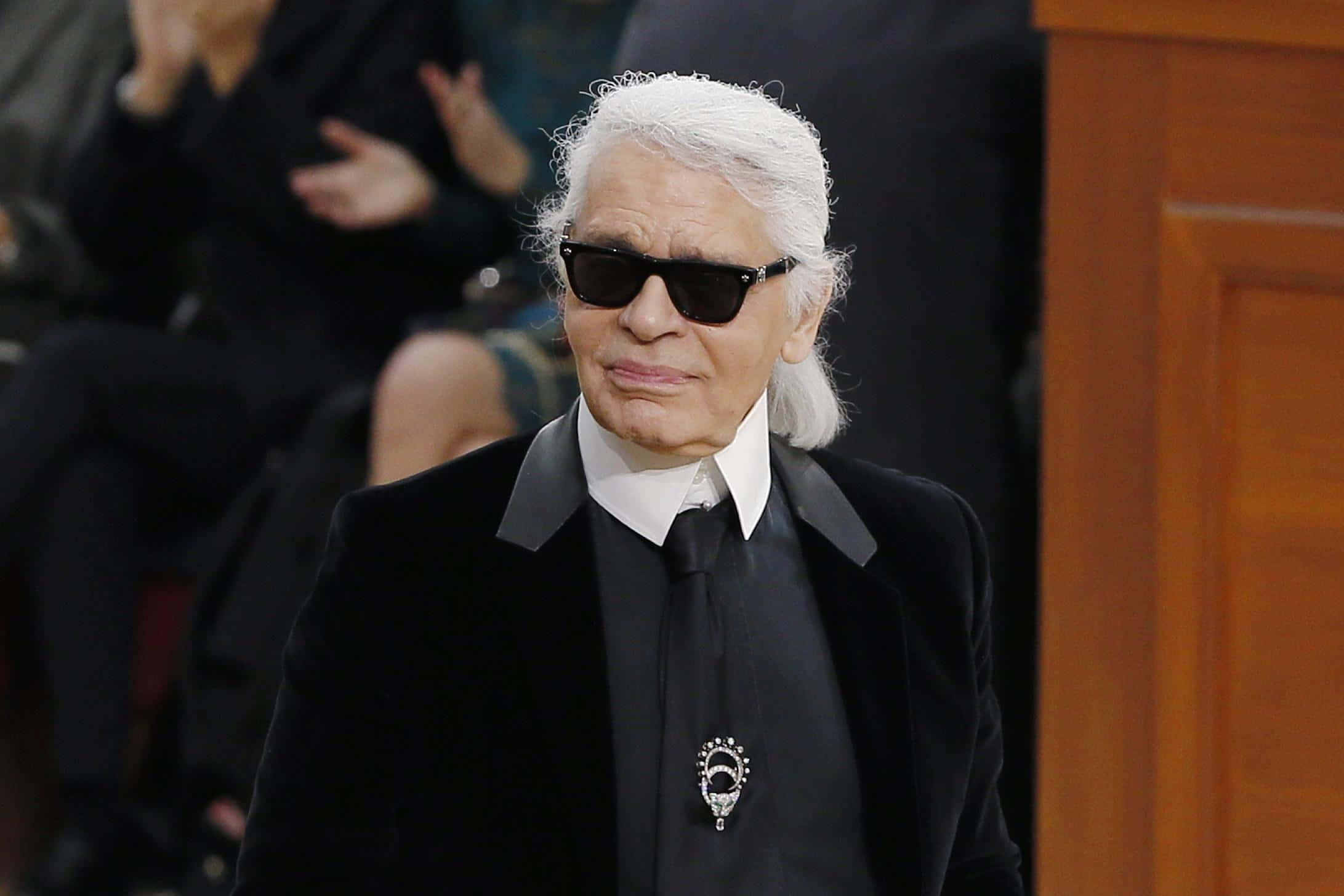 時裝設計師Karl Lagerfeld 壁紙 — 卡爾·拉格斐穿著黑色西裝和太陽眼鏡，表情嚴肅地走在時尚活動中。