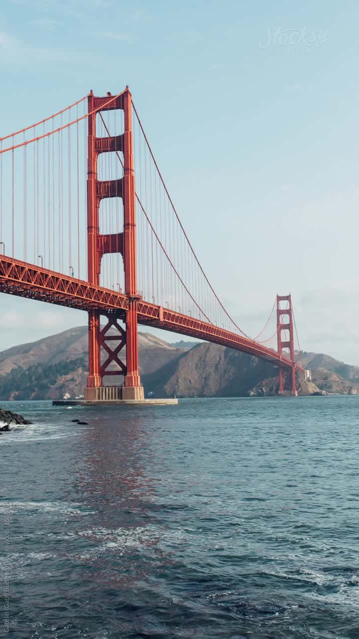 晨曦中的美麗石拱橋 壁紙 — 標誌性的紅色金門大橋橫跨在平靜的藍色海灣上，背景是朦朧的山丘，天空晴朗。