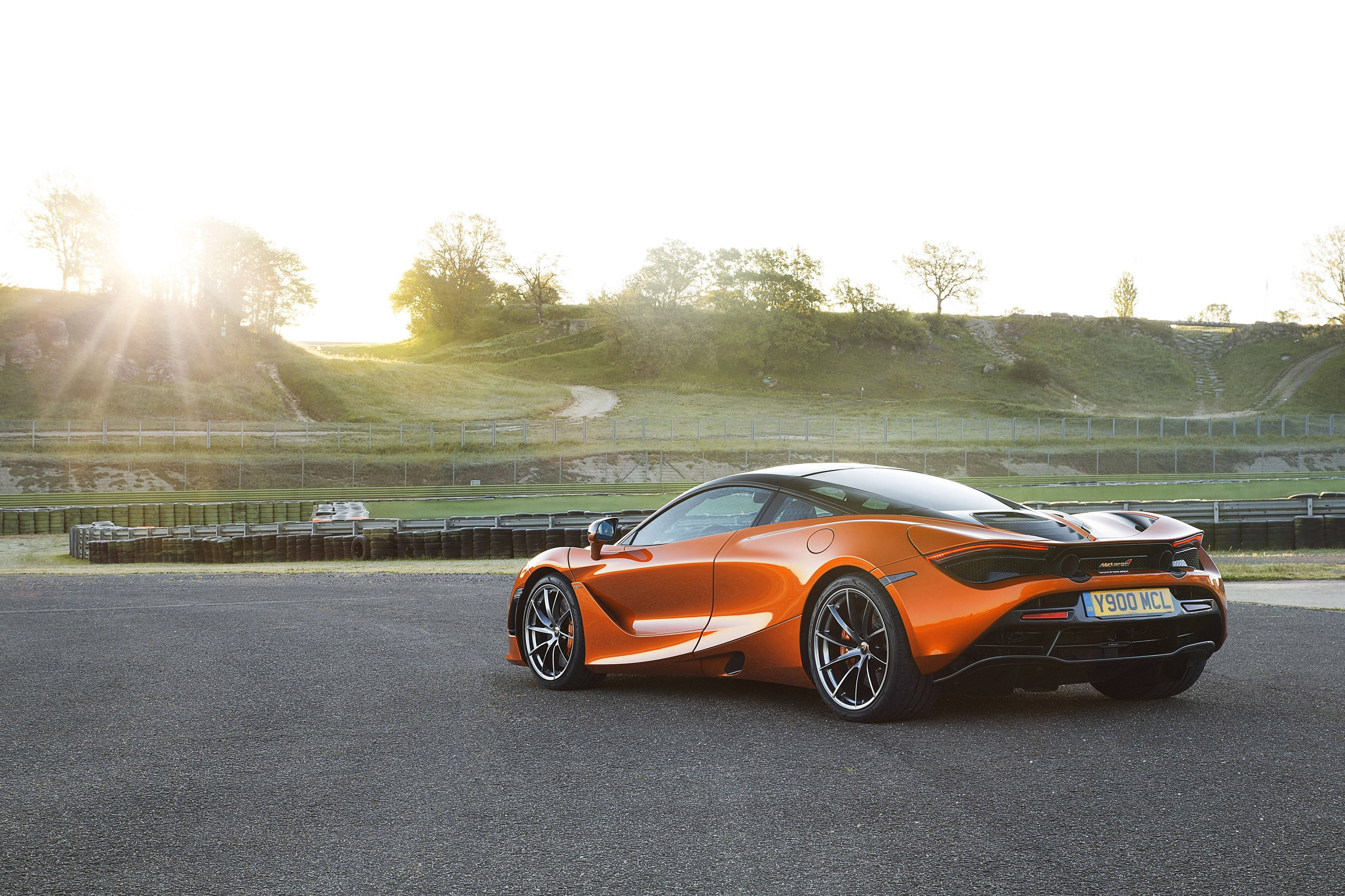 晨間駕駛麥拉倫720s 4K 壁紙 — 一輛充滿活力的橙色McLaren跑車在瀝青賽道上閃閃發光，金色的陽光穿過樹木灑在起伏的綠色山坡上。