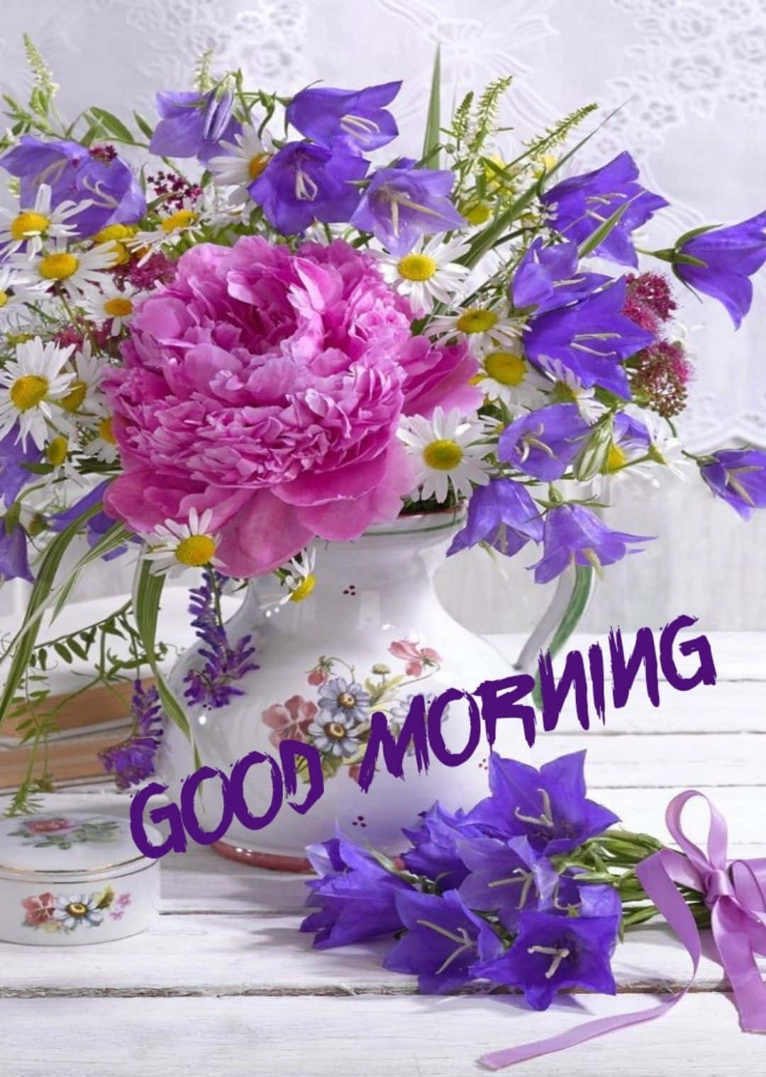 粉紅色牡丹和紫色花朵放在一個花卉圖案的花瓶中，帶有'GOOD MORNING'文字，置於白色木質表面上。