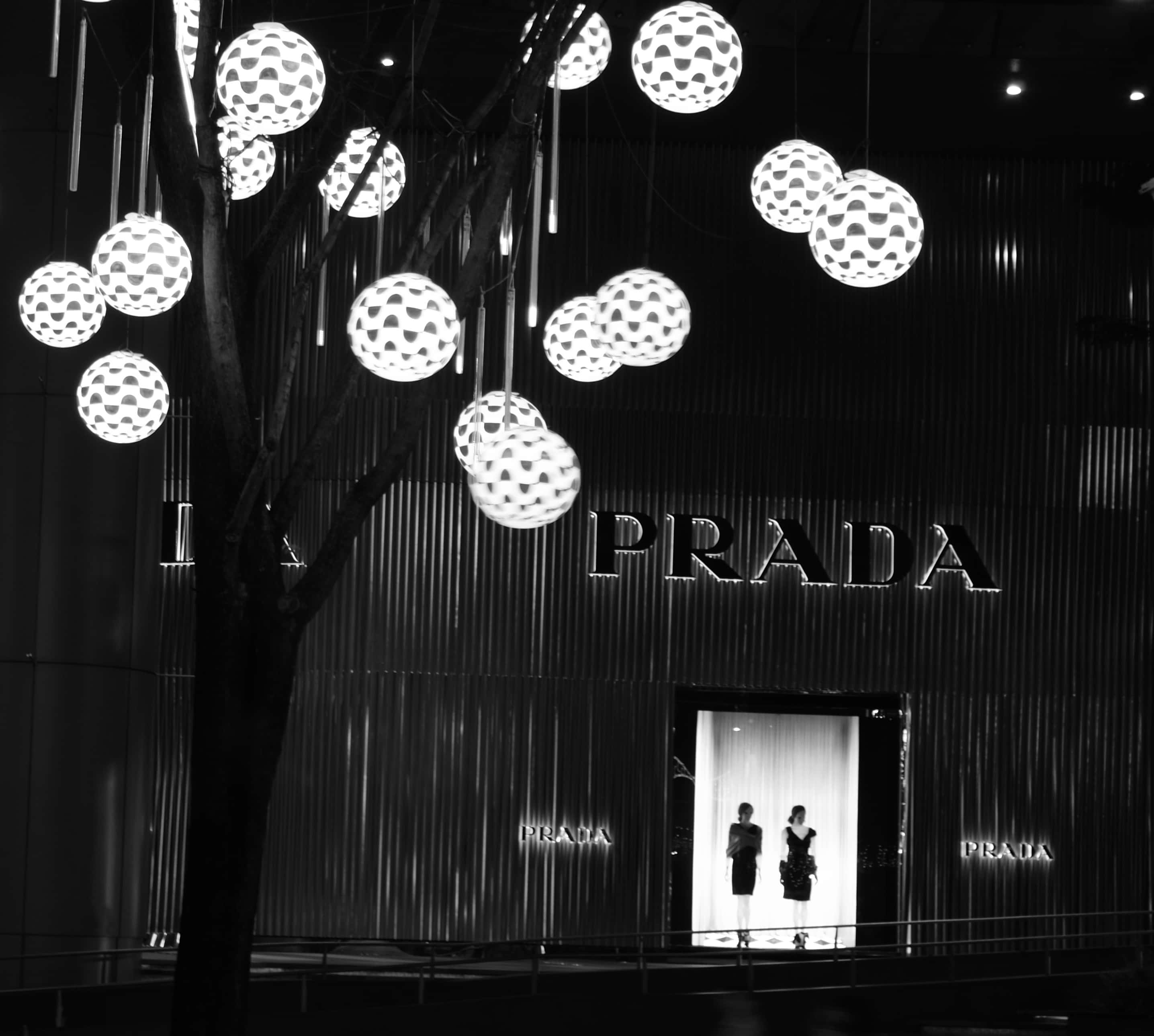 普拉達經典手袋：優雅永恆 壁紙 — 一個黑白城市夜景， featuring Prada 商店，掛有球形燈飾，前面有一棵樹。