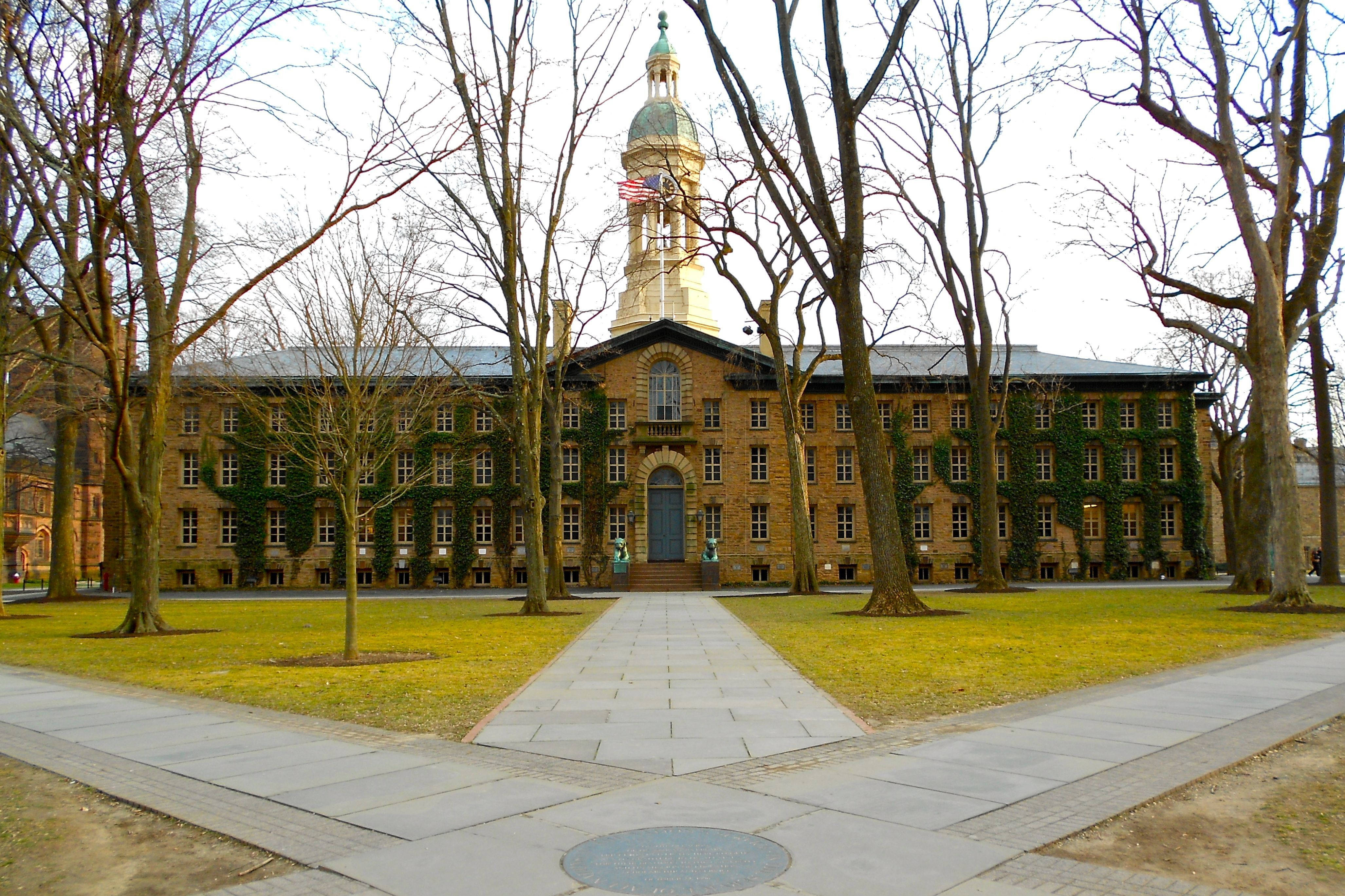 普林斯頓大學歷史性納薩大樓 壁紙 — 一座宏偉的歷史大學建築，中央有一座鐘樓，周圍是光禿禿的冬季樹木和一條通往拱形入口的鋪砌步道。