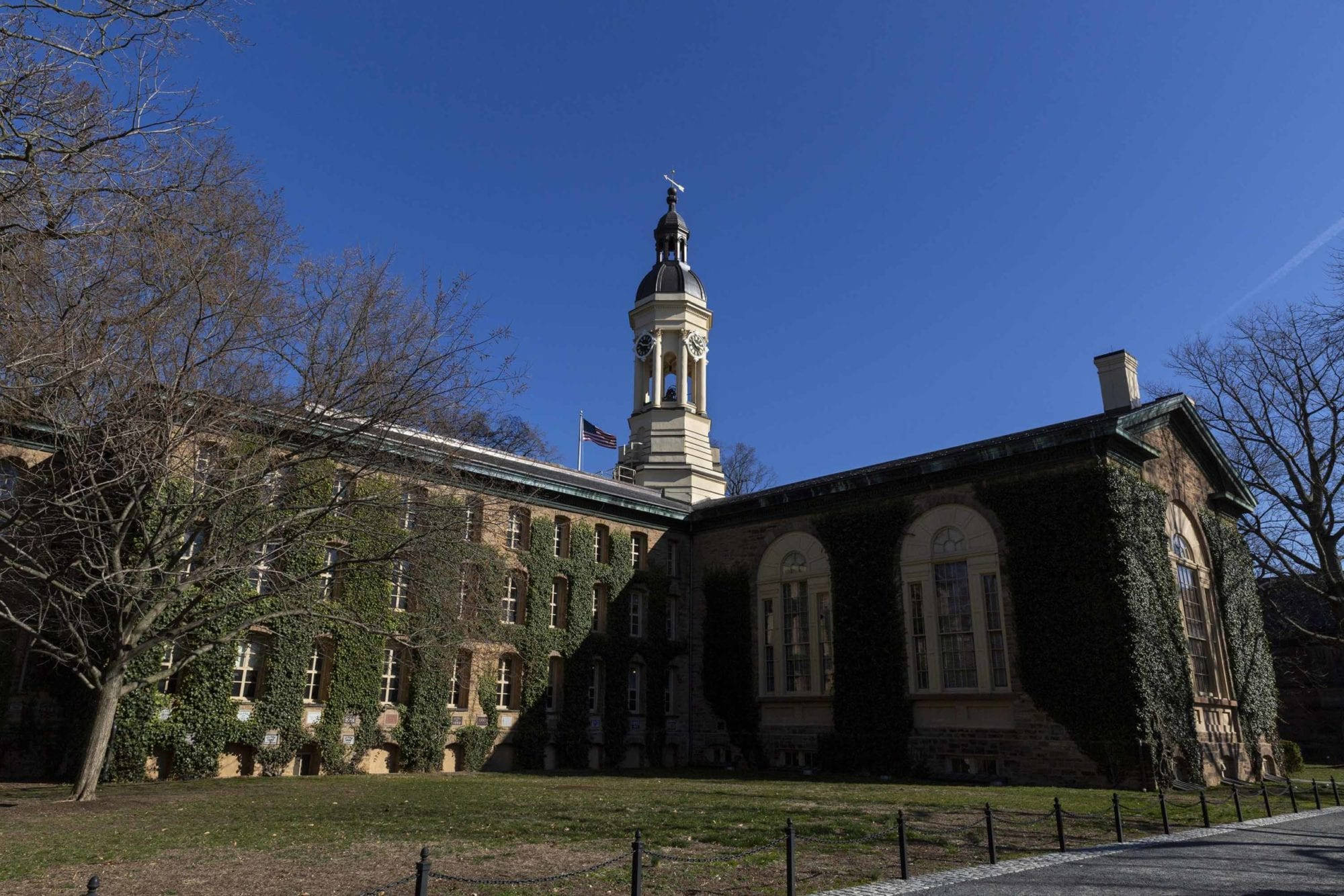 普林斯頓大學納薩廳 壁紙 — 一座宏偉的歷史石建築，帶有鐘樓和常春藤覆蓋的牆壁，矗立在晴朗的藍天下，樹木光禿。