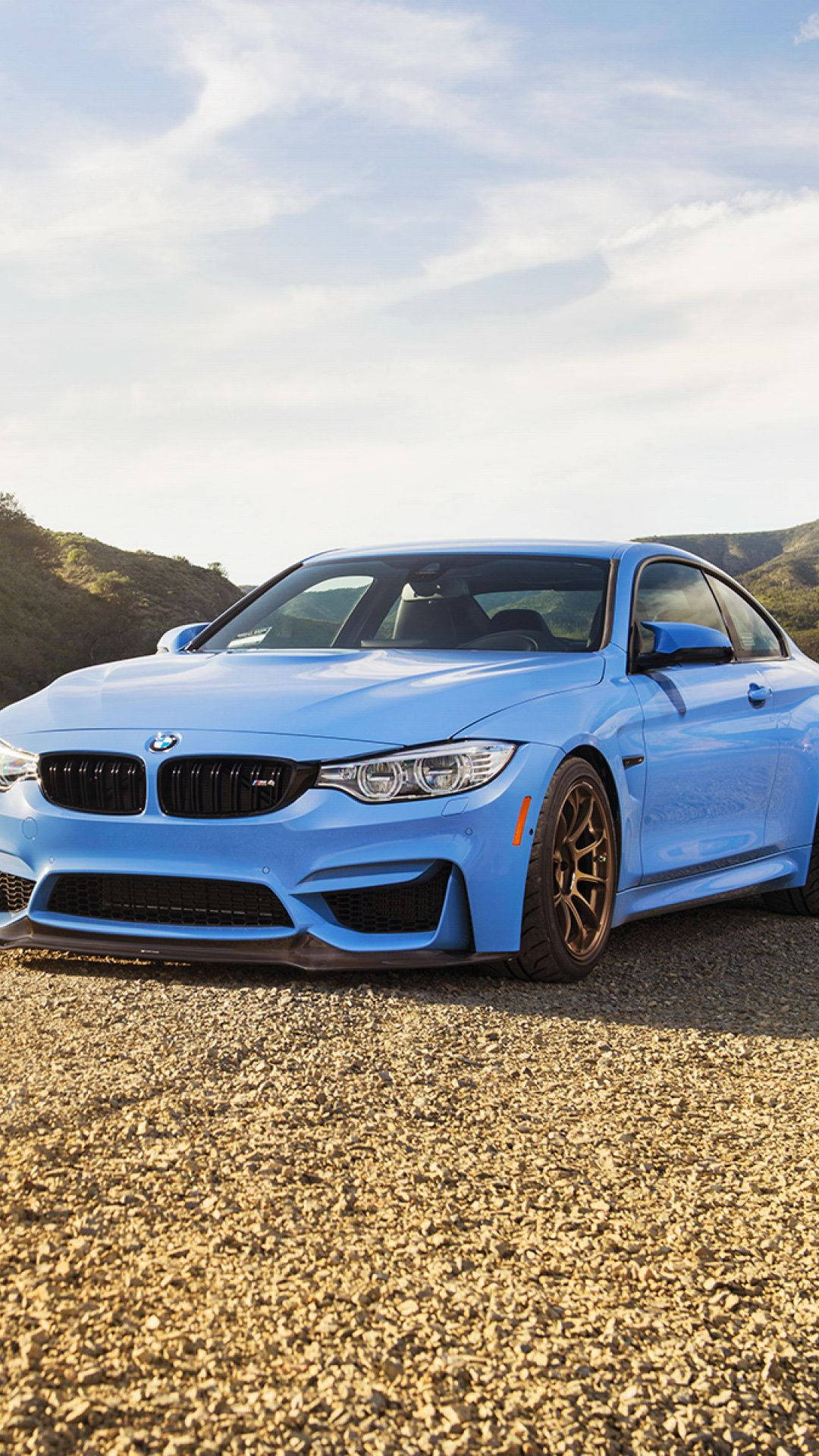 晴日下的嬰兒藍寶馬 壁紙 — 一輛充滿活力的藍色BMW M4雙門跑車停在碎石上，背景是起伏的丘陵，天空部分多雲。