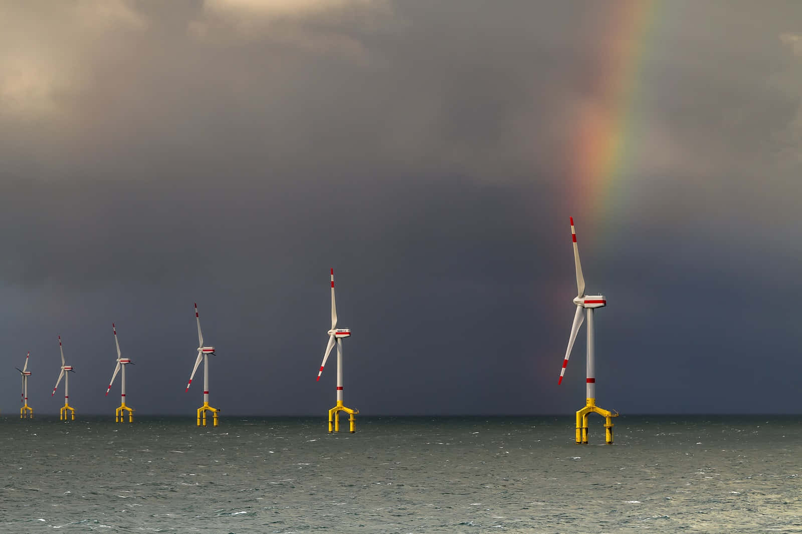 晴日海風輕拂的寧靜海灘 壁紙 — 一排離岸風力發電機矗立在波濤洶湧的灰色海水中，上方是暴風雨的天空，被一道鮮豔的彩虹穿透。