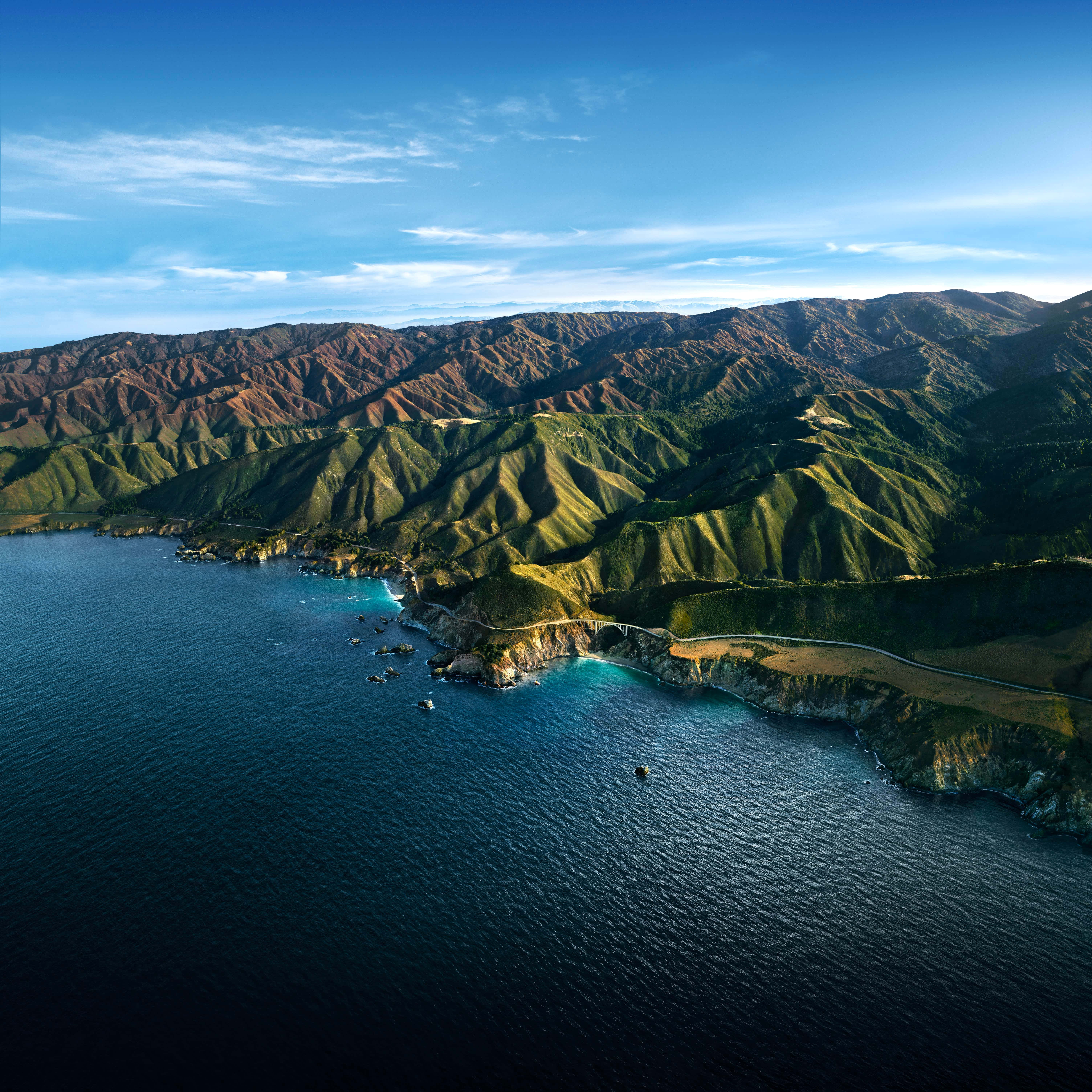 Big Sur 桌布 壁紙 — 晴朗天空下，崎嶇海岸山脈的鳥瞰圖，蜿蜒的道路緊貼懸崖，襯托著深藍色的海洋。