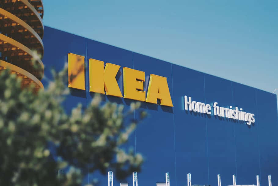 Ikea 桌布 壁紙 — 晴朗藍天下，明亮的藍色IKEA商店外牆配以醒目的黃色標誌，前景有模糊的綠色樹葉作框景。