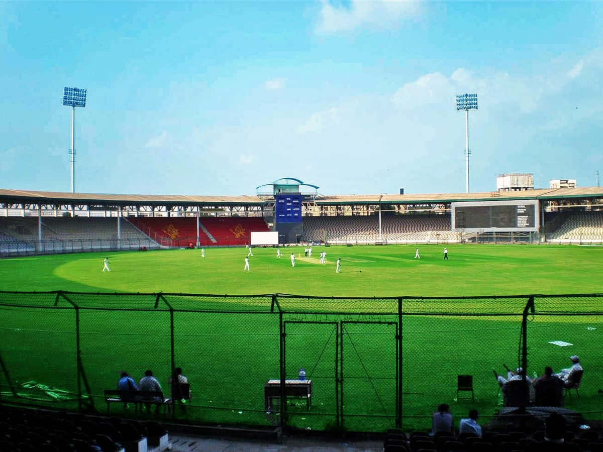 Cricket Ground 桌布 壁紙 — 晴朗藍天下板球場的廣闊視野，球員在充滿活力的綠色場地上，觀眾坐在看台上。