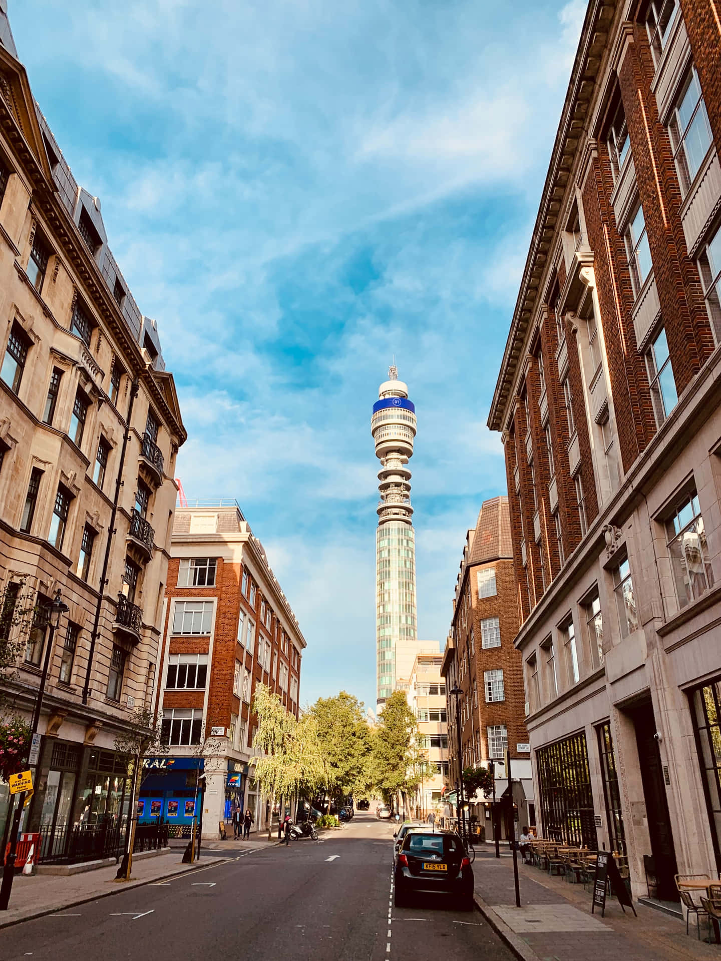 晴空下BT塔街景手機壁紙 壁紙 — 一條狹窄的倫敦街道，兩旁是歷史悠久的磚砌建築，向上延伸至標誌性的BT塔，刺破明亮的藍天。