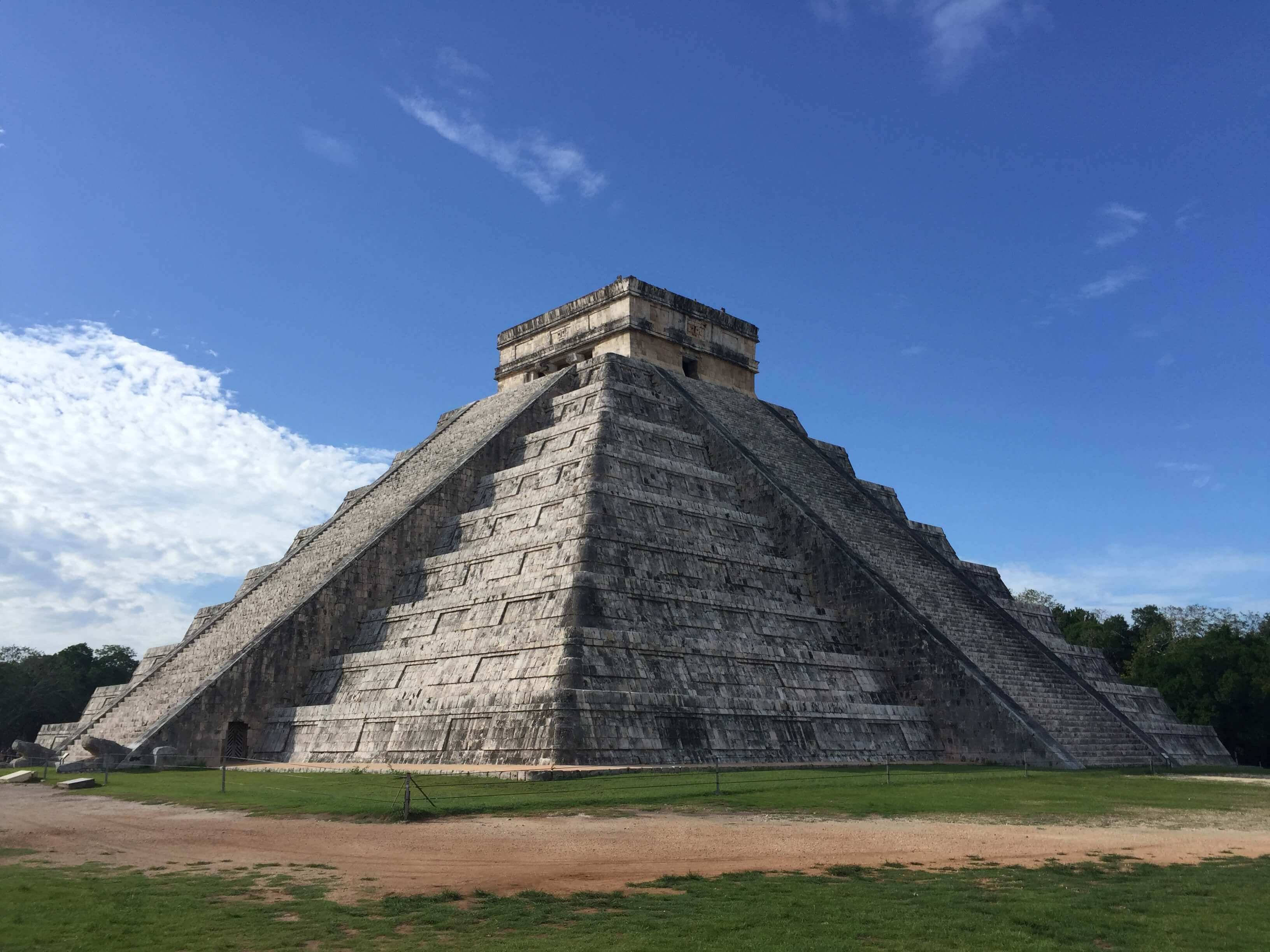 晴空下的奇琴伊察 壁紙 — 一座巨大的古瑪雅金字塔在明亮的藍天下升起，天空有散落的雲朵，底部有綠草。